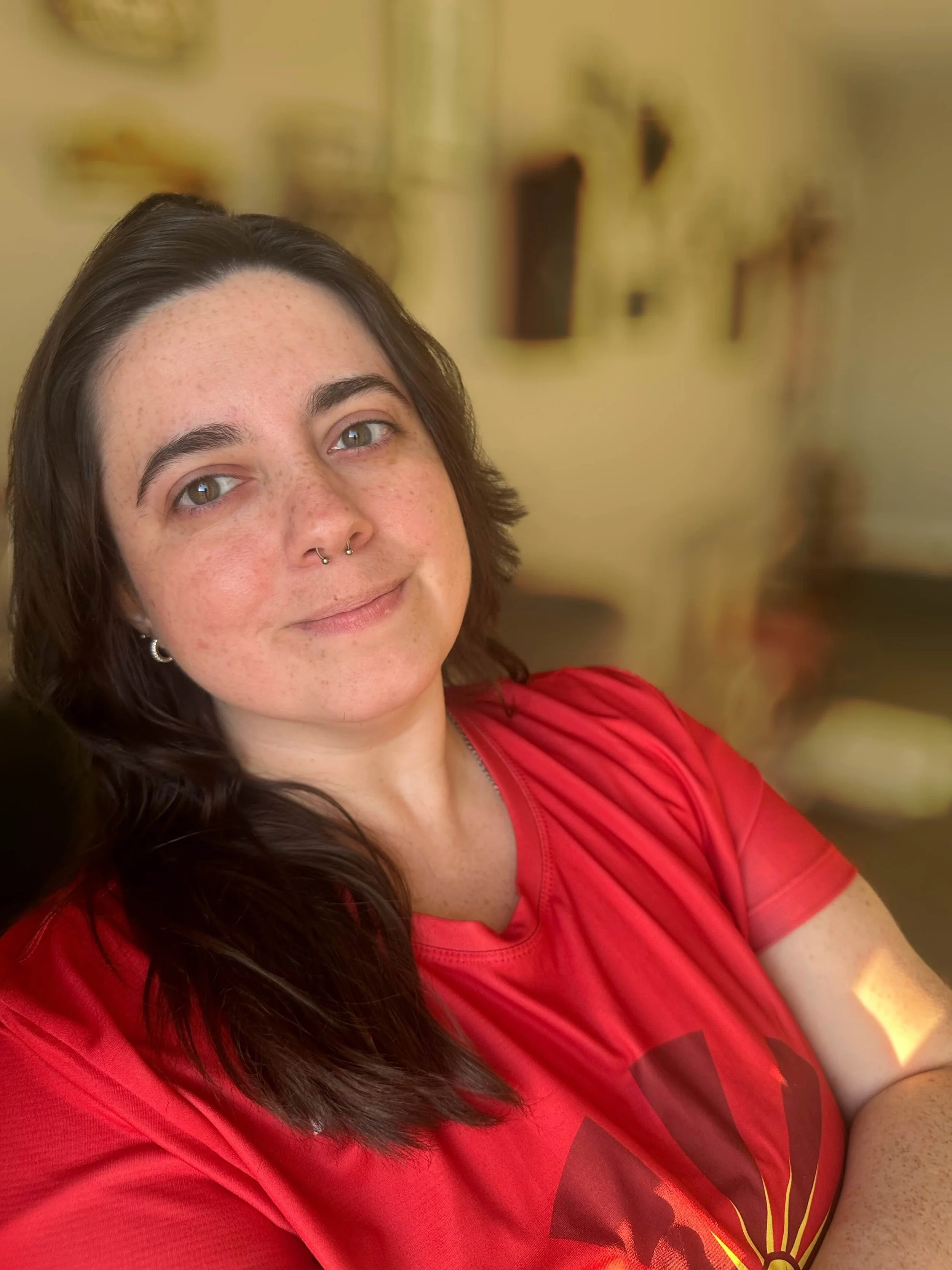 A woman with dark brown hair, hazel eyes, and freckles wearing a septum piercing and a red T-shirt.
