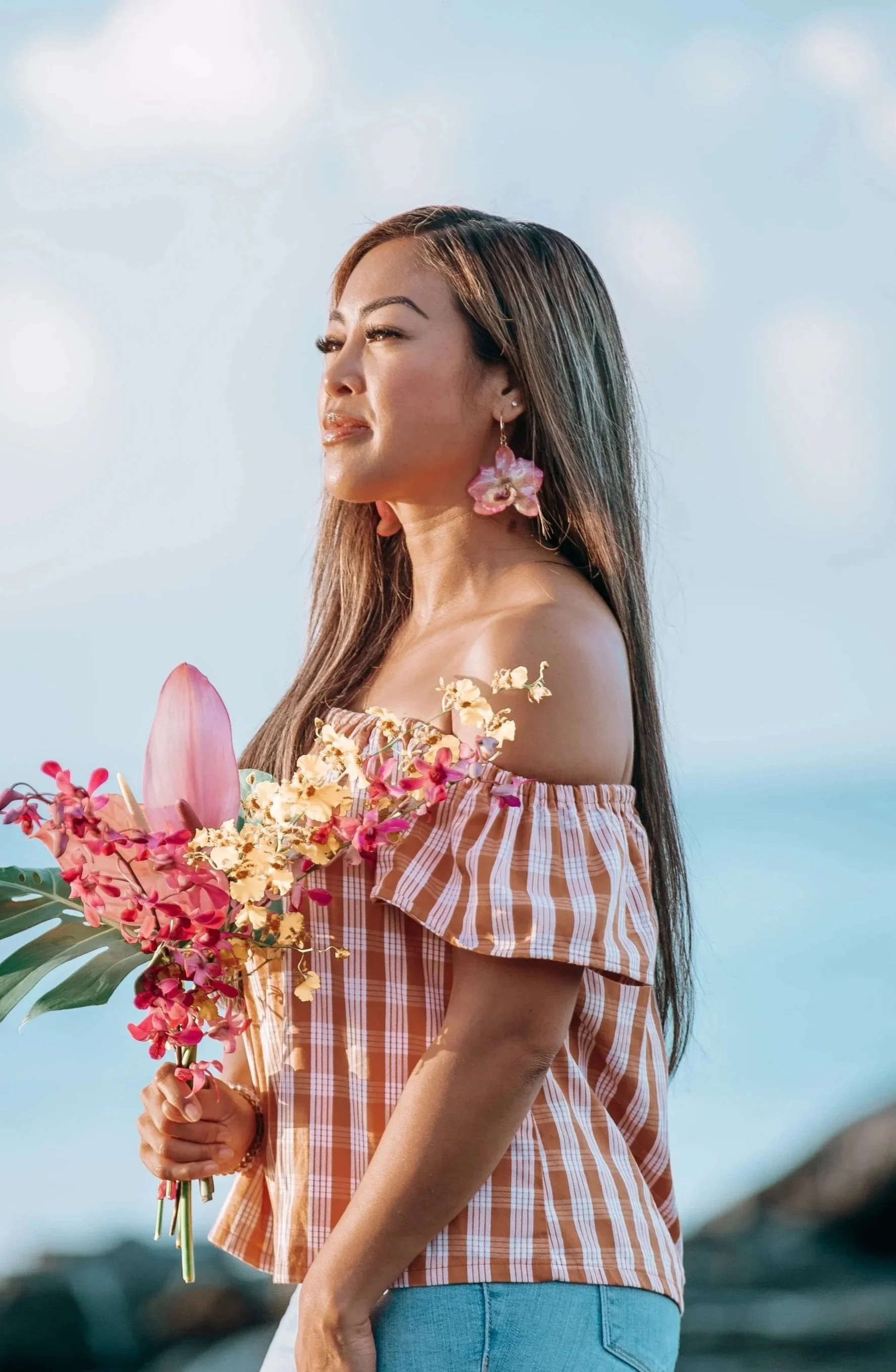 Outdoor portrait of a woman holding pink and yellow tropical flowers against a soft island sky during a Flora & Femina session in Honolulu.