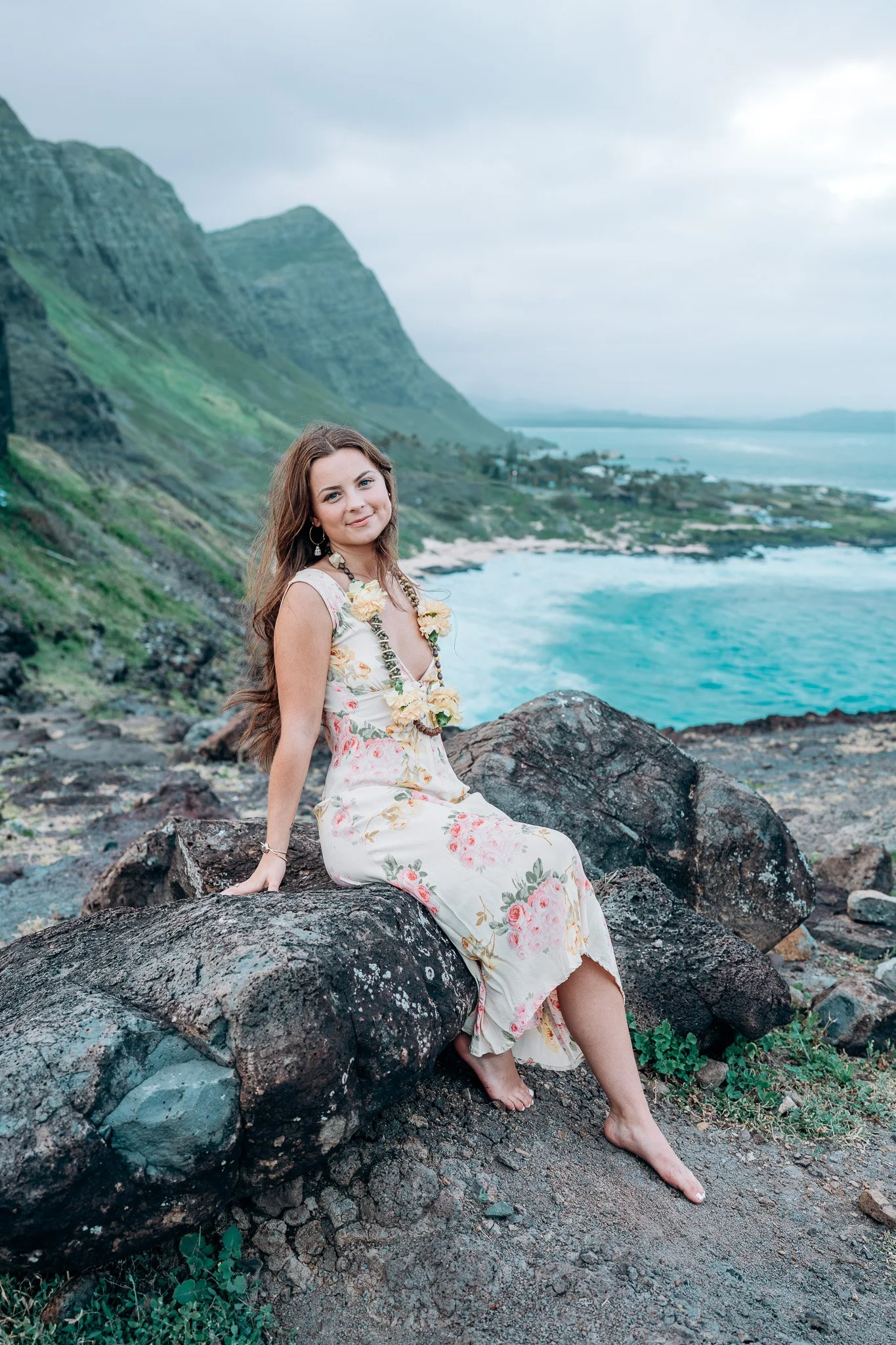 Outdoor coastal portrait of a woman sitting on rocks by the ocean with green Oʻahu mountains in the background during a Flora & Femina session.