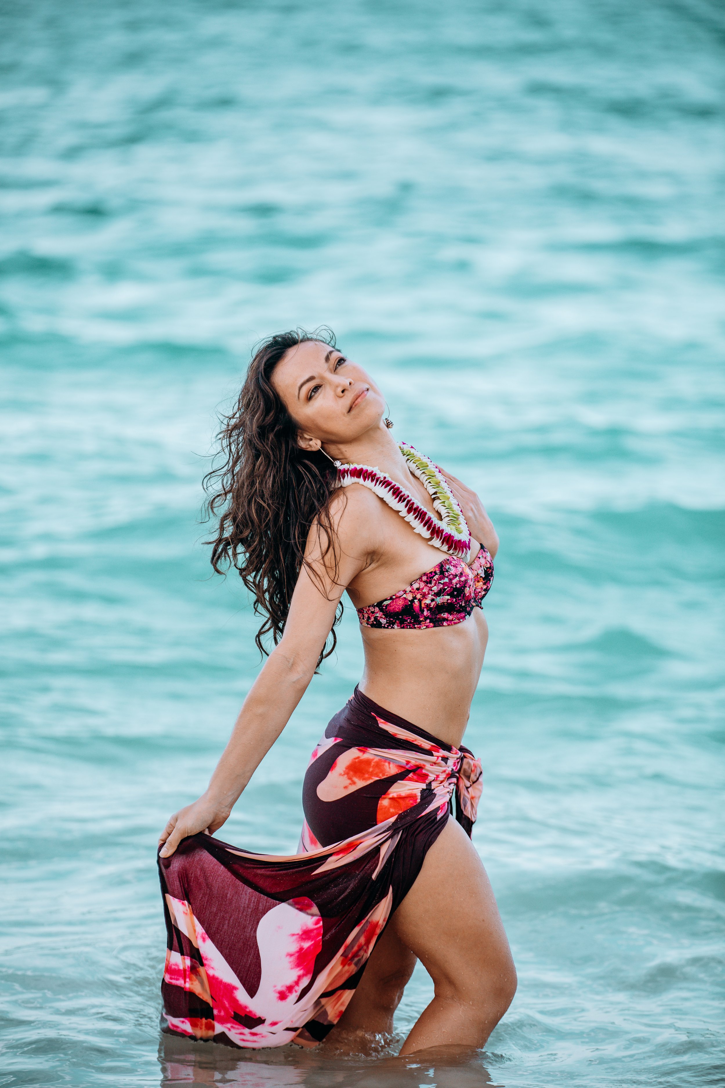 Beach portrait of a woman wearing a colorful bikini and floral sarong standing in the ocean with waves in the background.