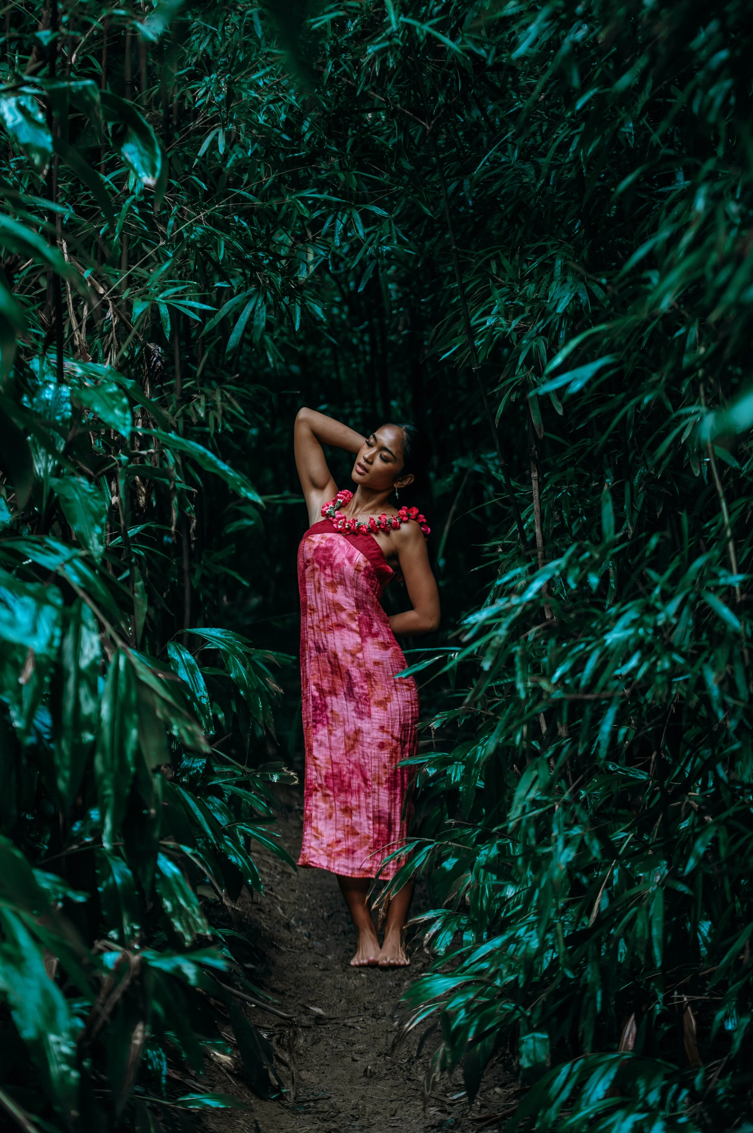 Outdoor portrait of a woman in a pink dress wearing a floral necklace on a lush green forest trail during a Flora & Femina session in Oʻahu.