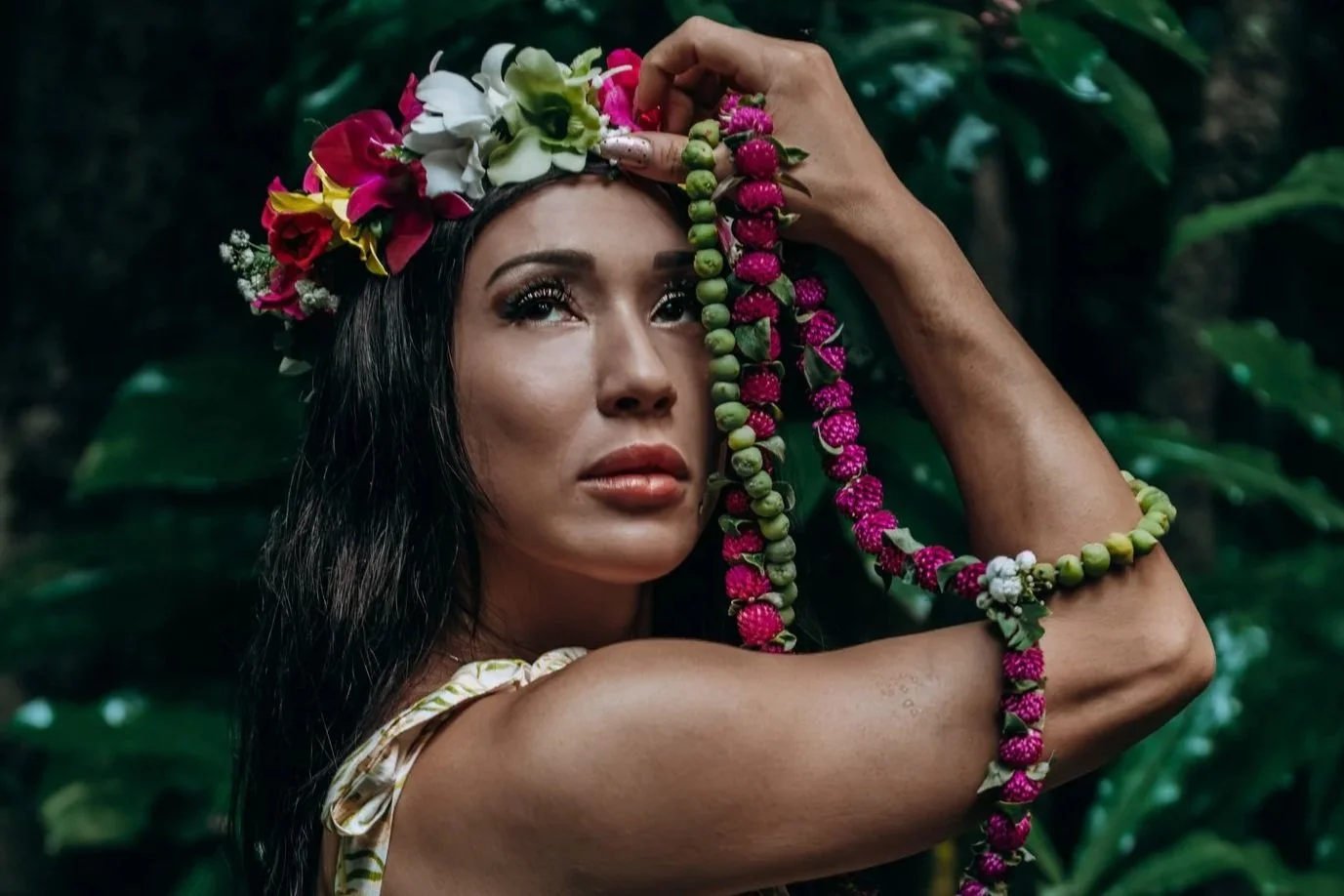 Tropical floral portrait of a woman wearing a flower crown and holding a beaded garland in a lush green jungle setting.