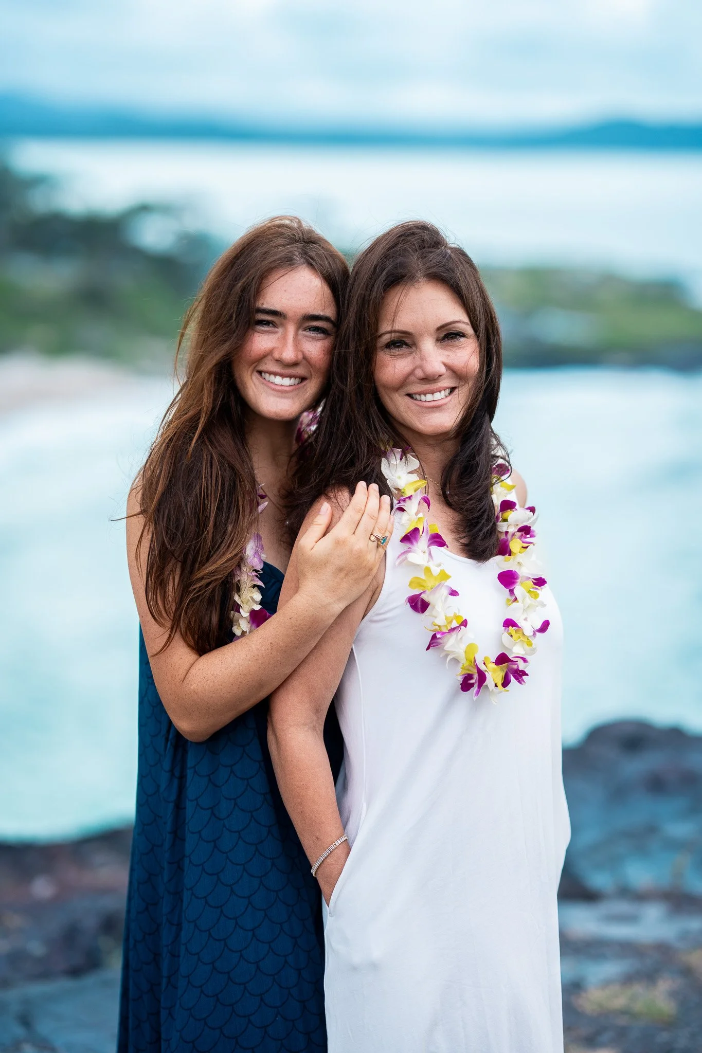 Beach portrait of two women wearing Hawaiian flower leis with ocean waves in the background during an outdoor Flora & Femina session.