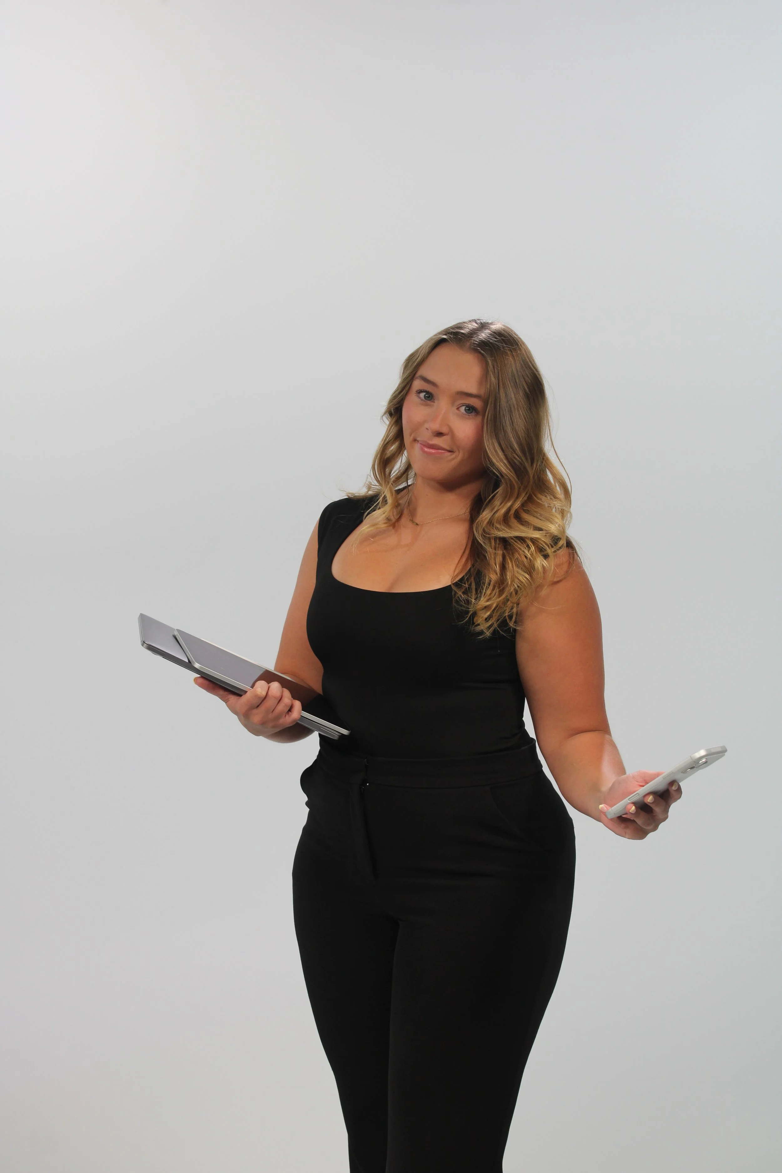 A young woman with wavy, blonde hair, wearing a black sleeveless top and black pants, holding a tablet in her left hand and a smartphone in her right hand, standing against a plain white background.
