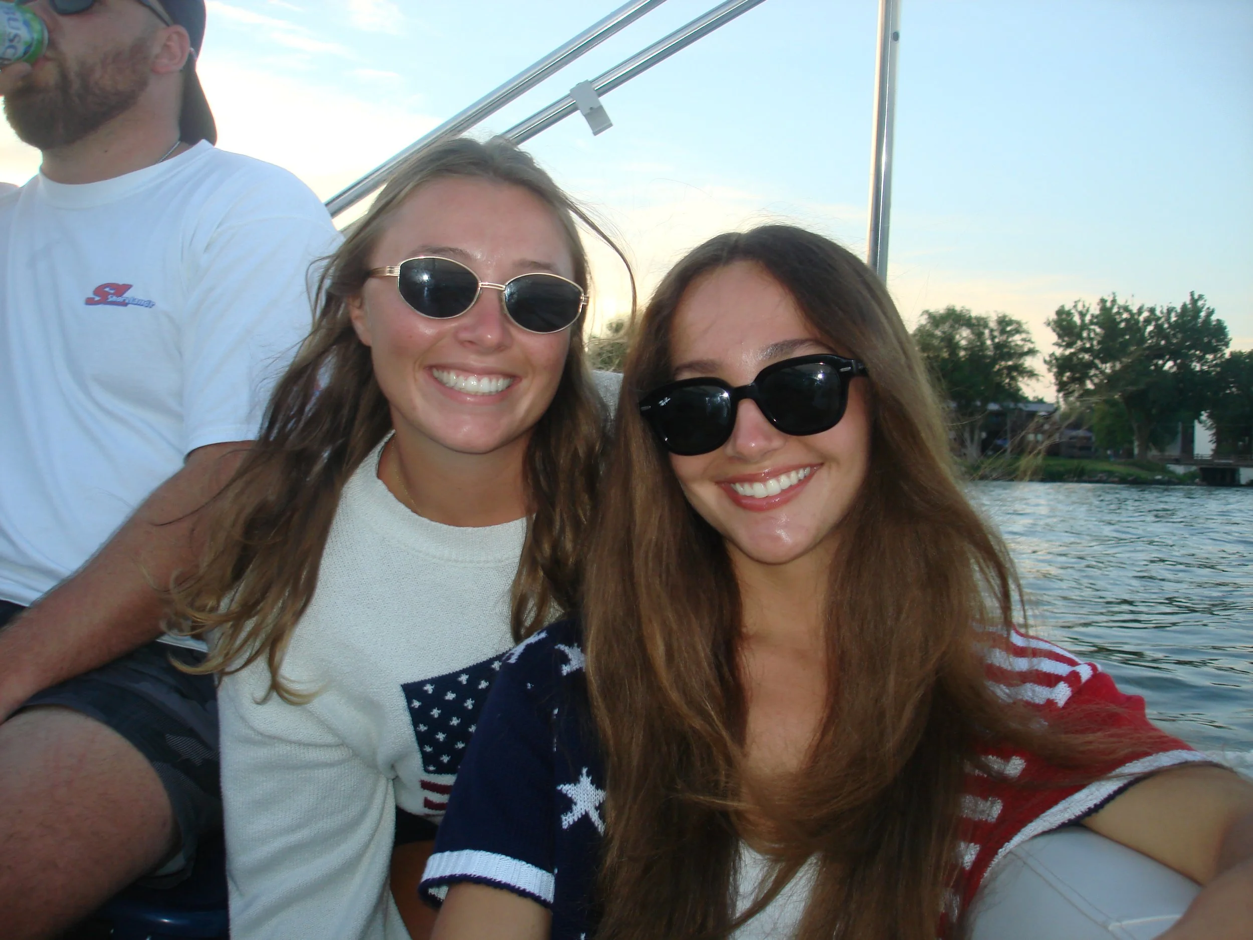 Two women smiling and wearing sunglasses in a boat near water with trees in the background.