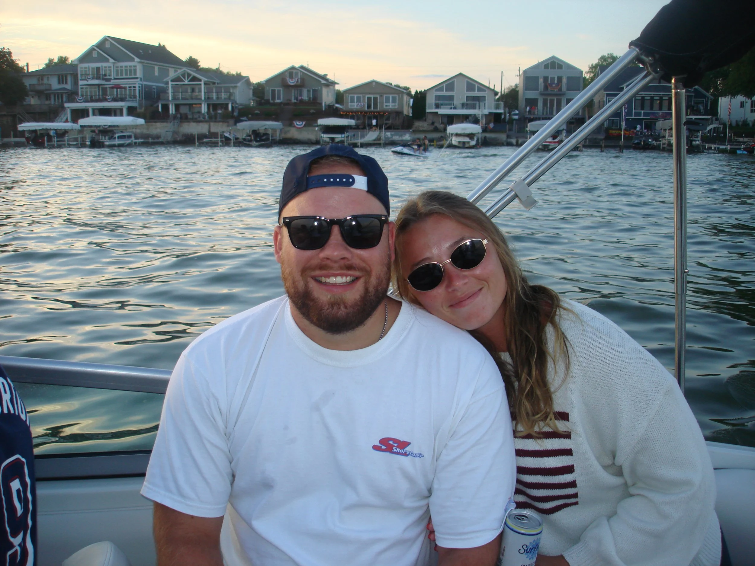 A smiling man with a beard, wearing sunglasses, a backward cap, and a white T-shirt, sitting close to a woman with blonde hair and black sunglasses, on a boat by a waterfront with houses and boats.