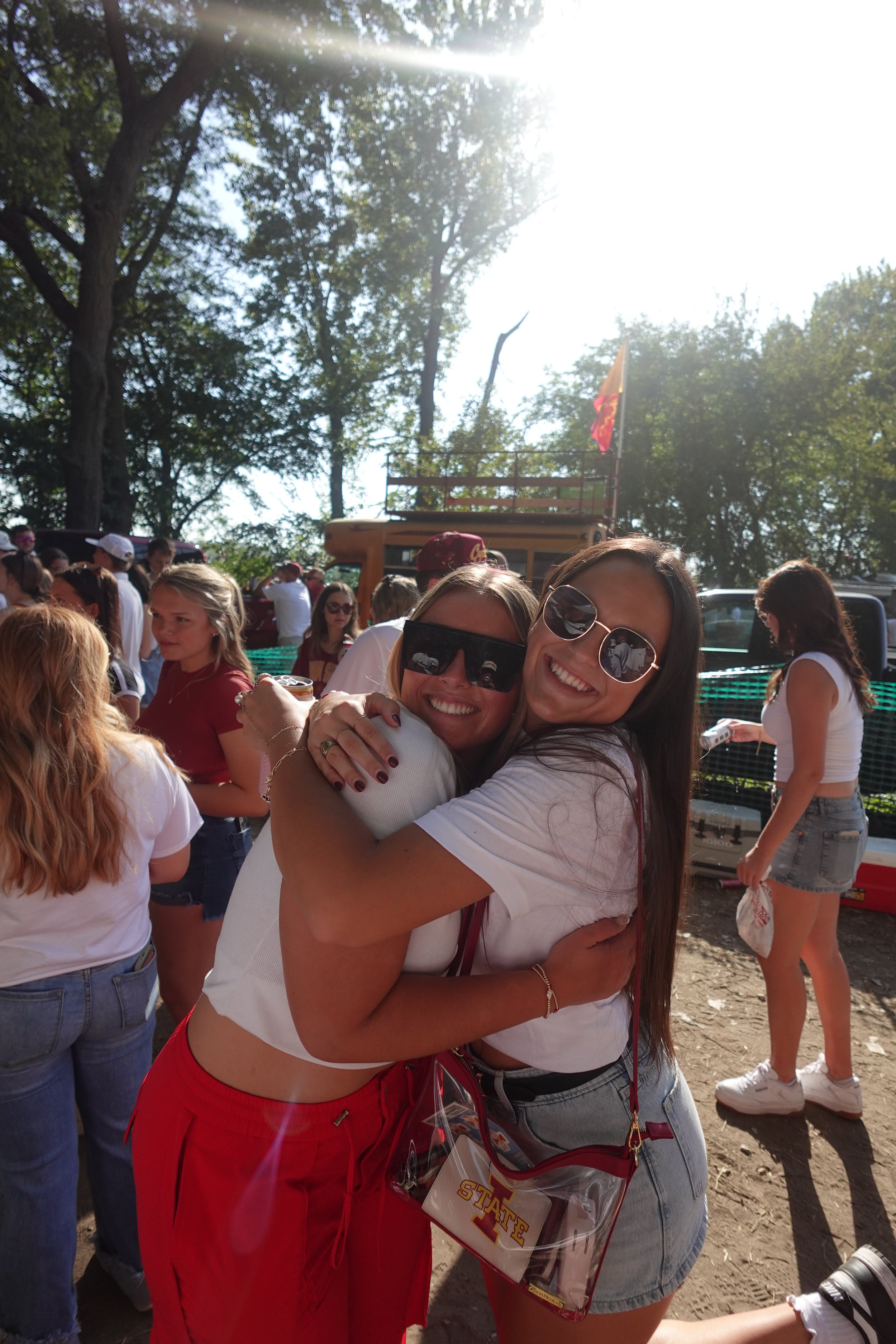 Two women hugging and smiling at an outdoor event, surrounded by a crowd of people, with trees and a sunny sky in the background.