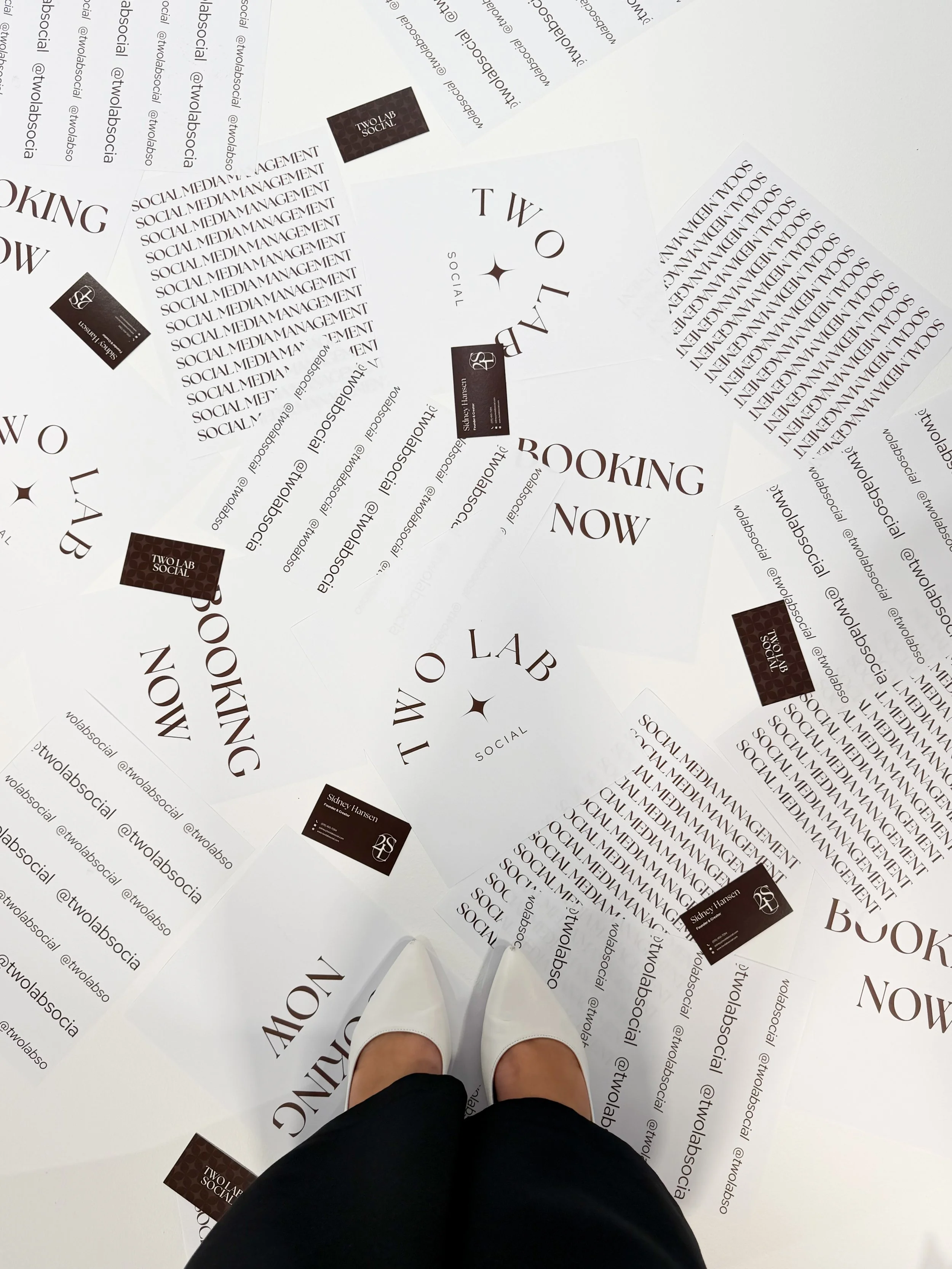 Photo of a person standing on a white floor covered with promotional flyers for Two Lab Social, with their feet visible at the bottom of the image. The flyers display the business name and social media handles.