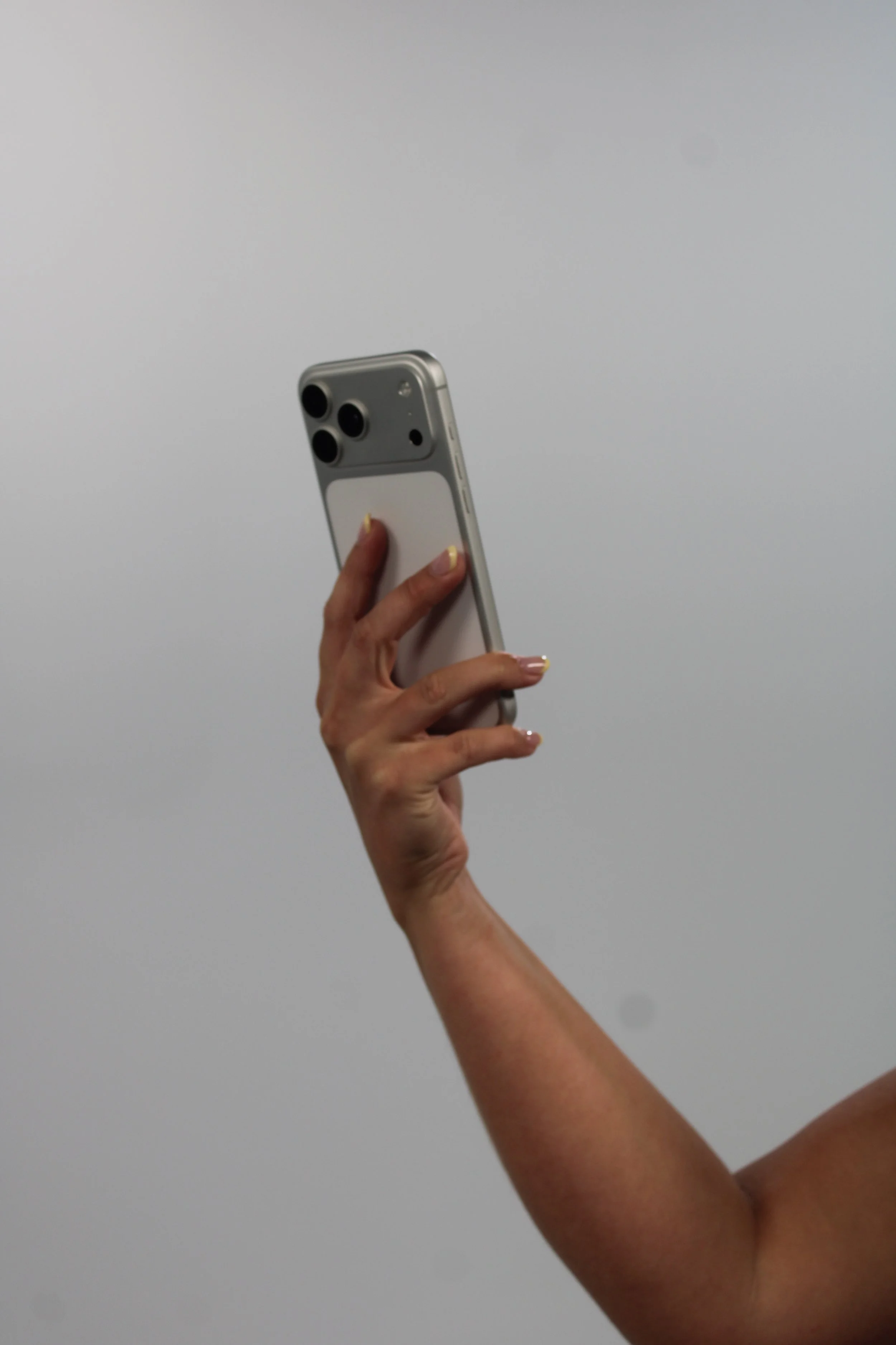 A person's hand holding a silver iPhone with a triple-lens camera, against a plain gray background.
