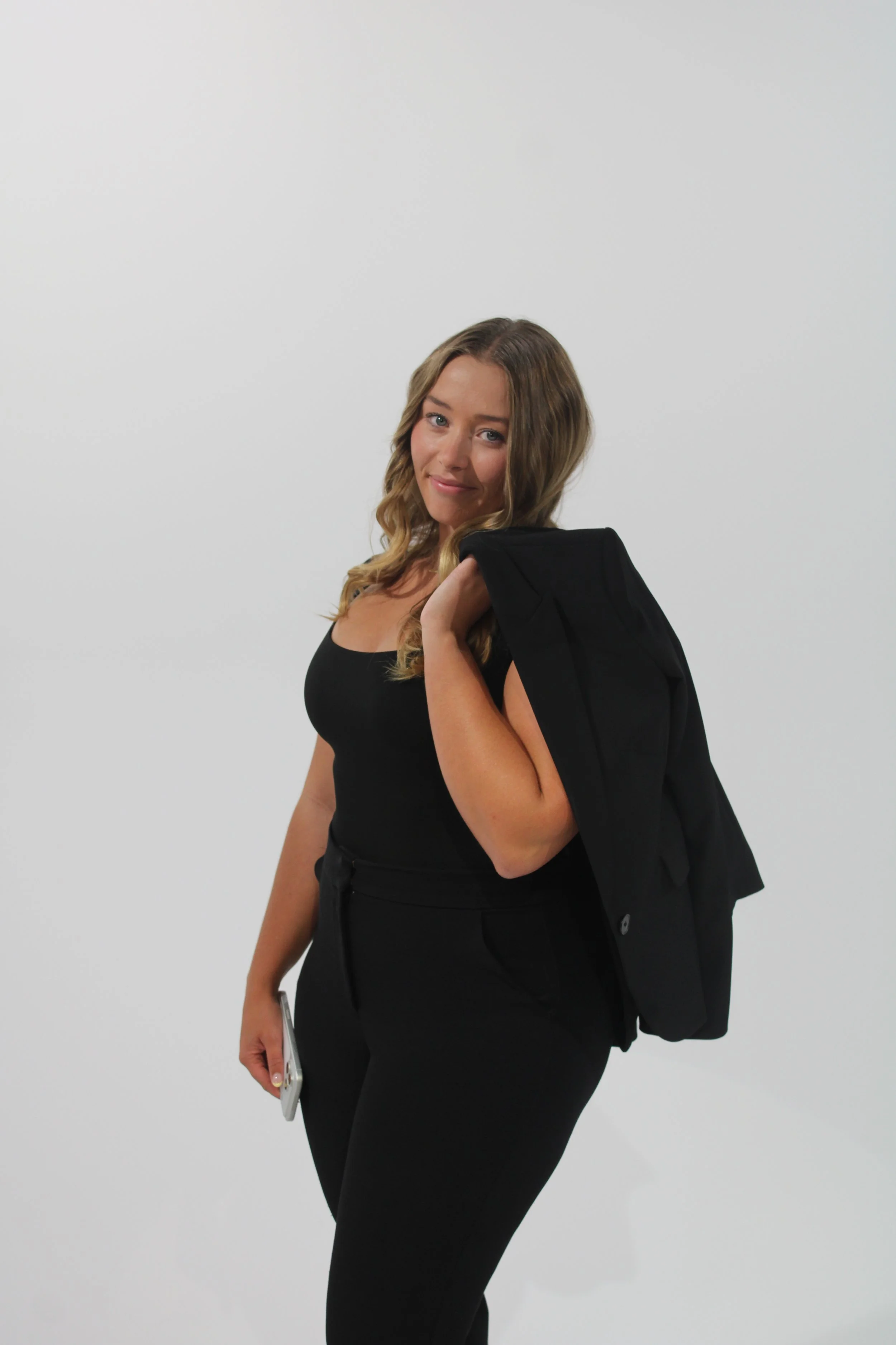 A woman with wavy light brown hair in a black sleeveless top and black pants, holding a smartphone in her right hand, is smiling slightly and looking at the camera. She has a black jacket draped over her left shoulder. The background is plain white.