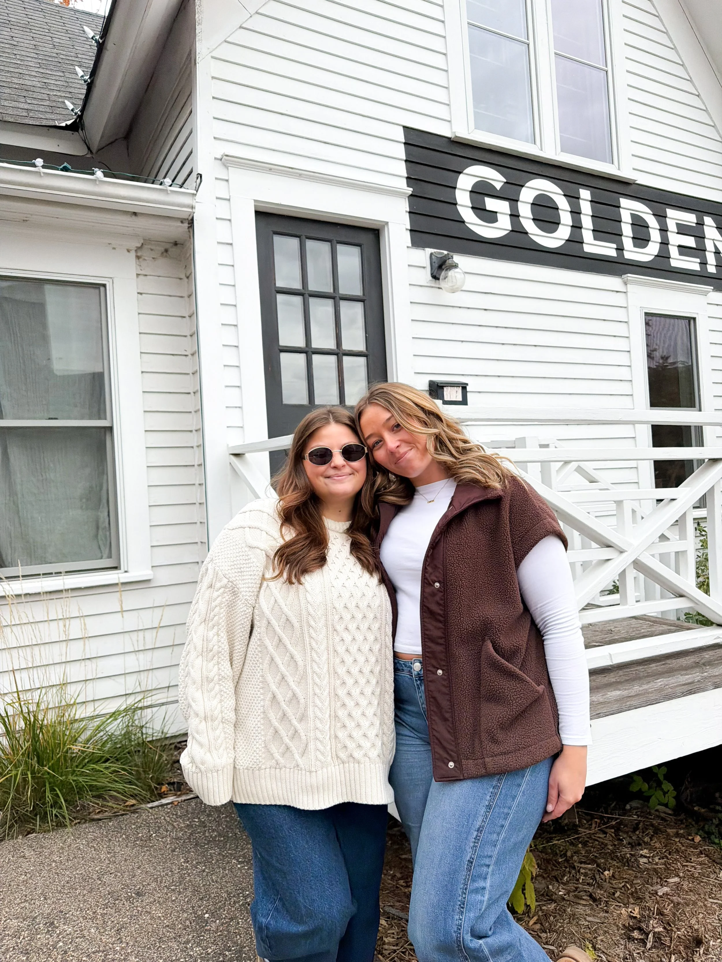Two women posing outdoors in front of a white house with the word "GOLDEN" on the house wall. One woman wears sunglasses and a cream sweater, while the other wears a white shirt and a brown vest. There are plants and a walkway in the foreground.