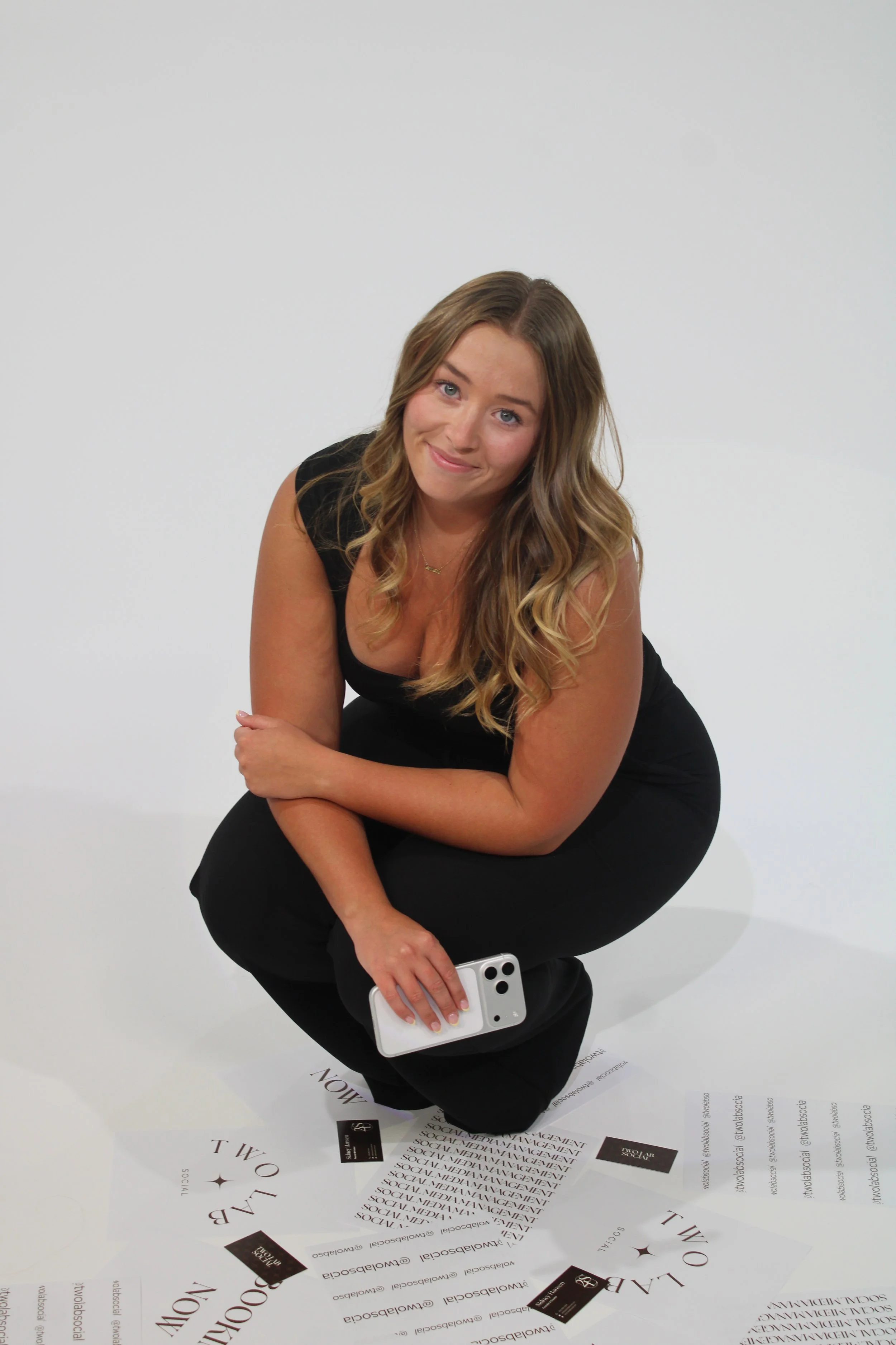 Woman with long wavy brown hair, wearing a black sleeveless top, squatting and smiling at camera, holding a smartphone, against a plain white background with scattered black and white Two Lab Social flyers on the floor.