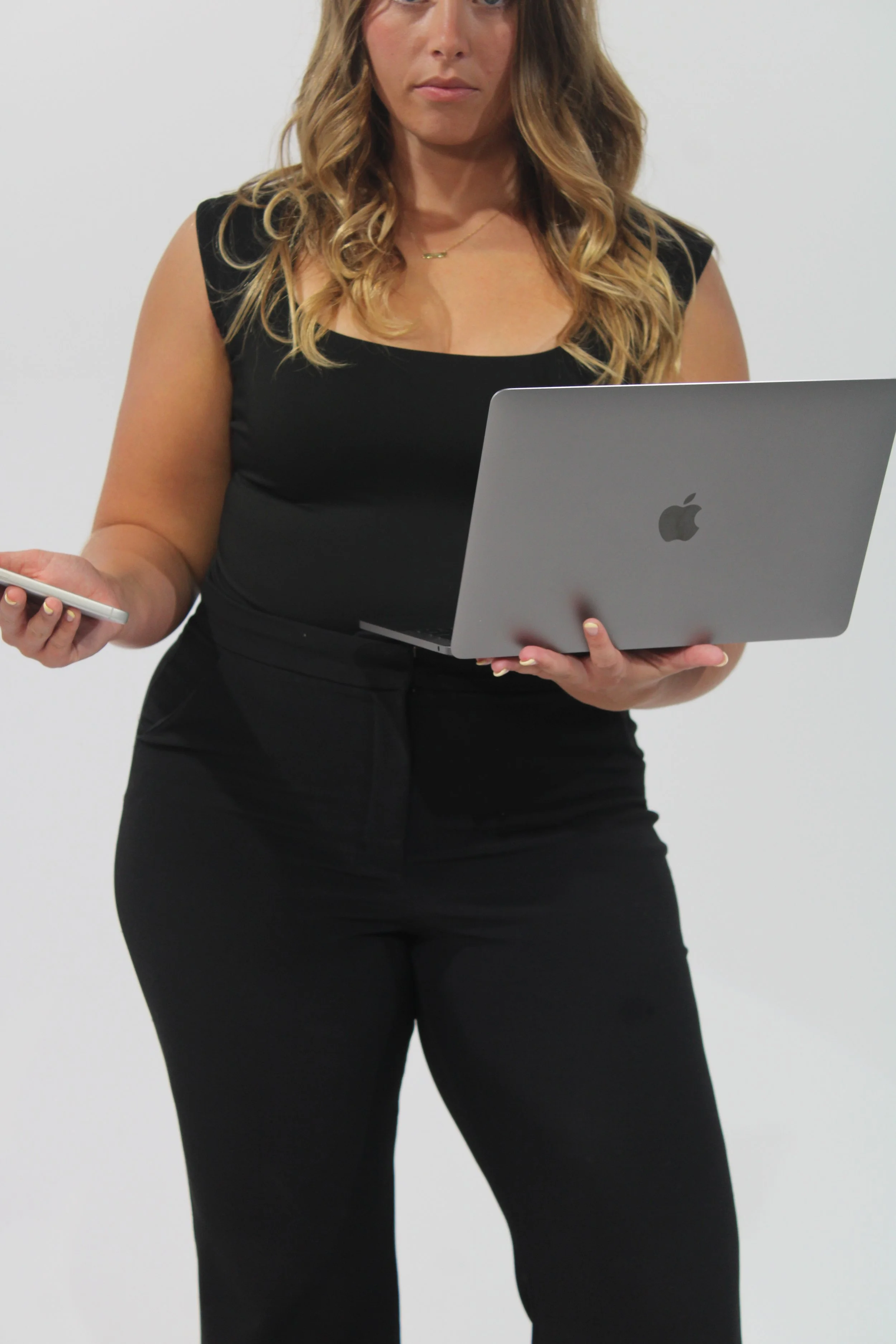 A woman with long wavy blonde hair wearing a black sleeveless top and black pants, holding a silver laptop and a smartphone, standing against a plain white background.
