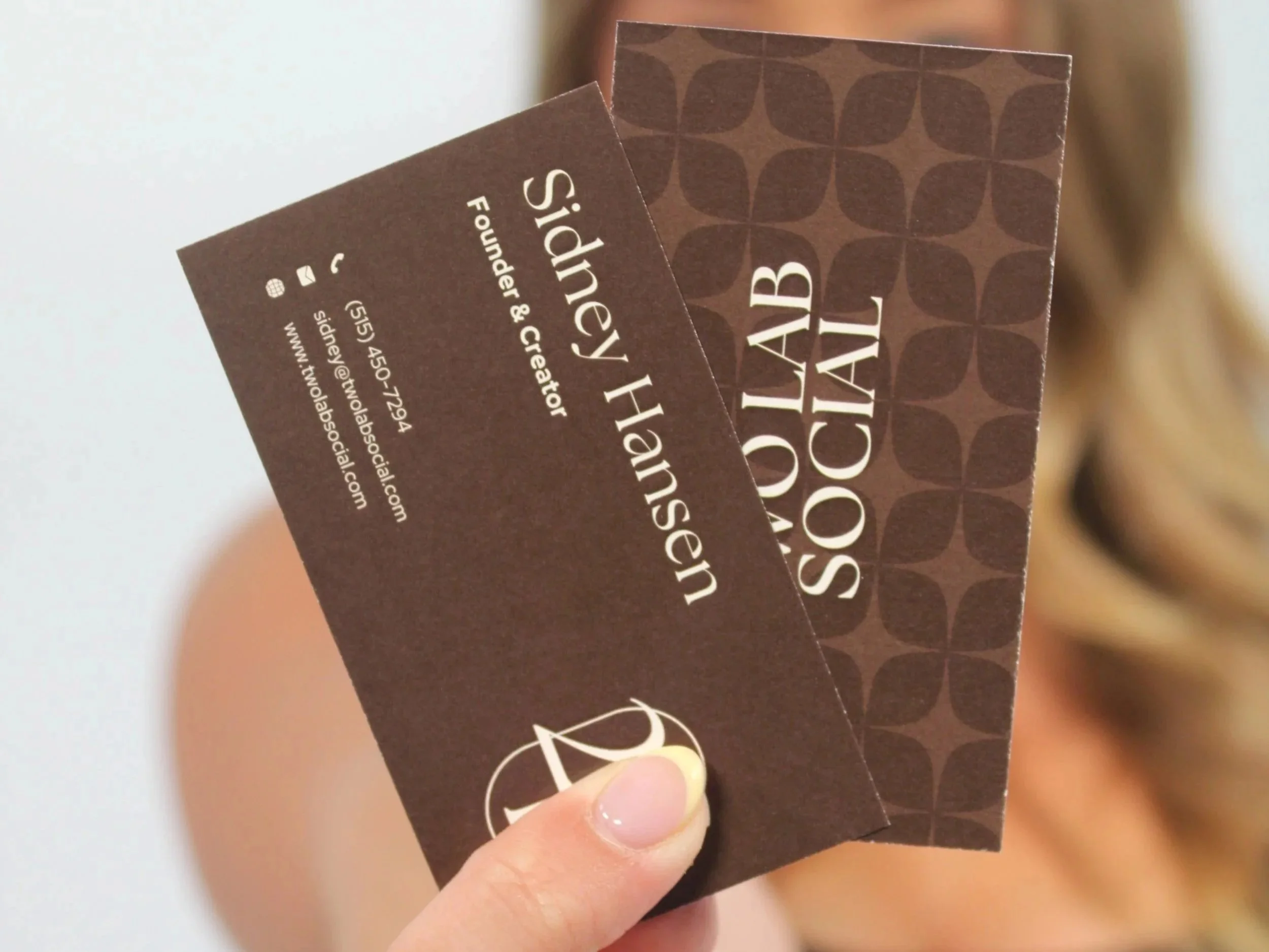 Person holding a Two Lab Social business card with dark brown background and patterned design, featuring logo and contact information in white text.