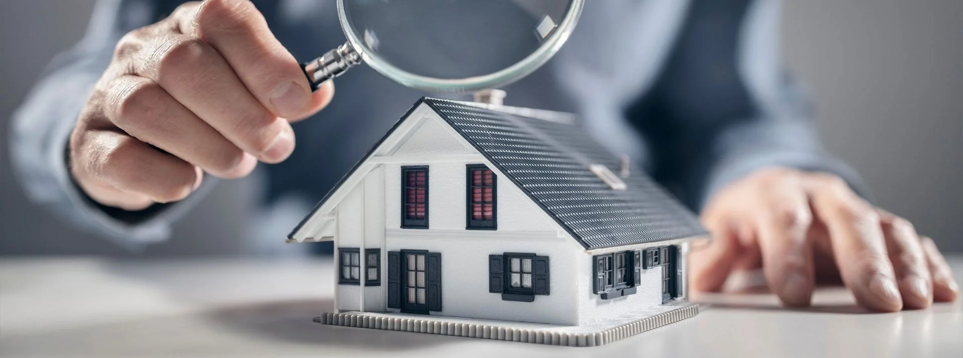 Person holding and inspecting a small model house with a magnifying glass.