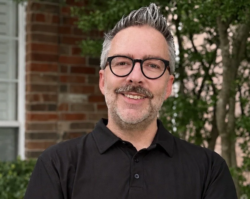 A professional man with brown and grey hair, glasses, and a moustache and five o'clock shadow, smiling outdoors in front of a house with bricks and trees.