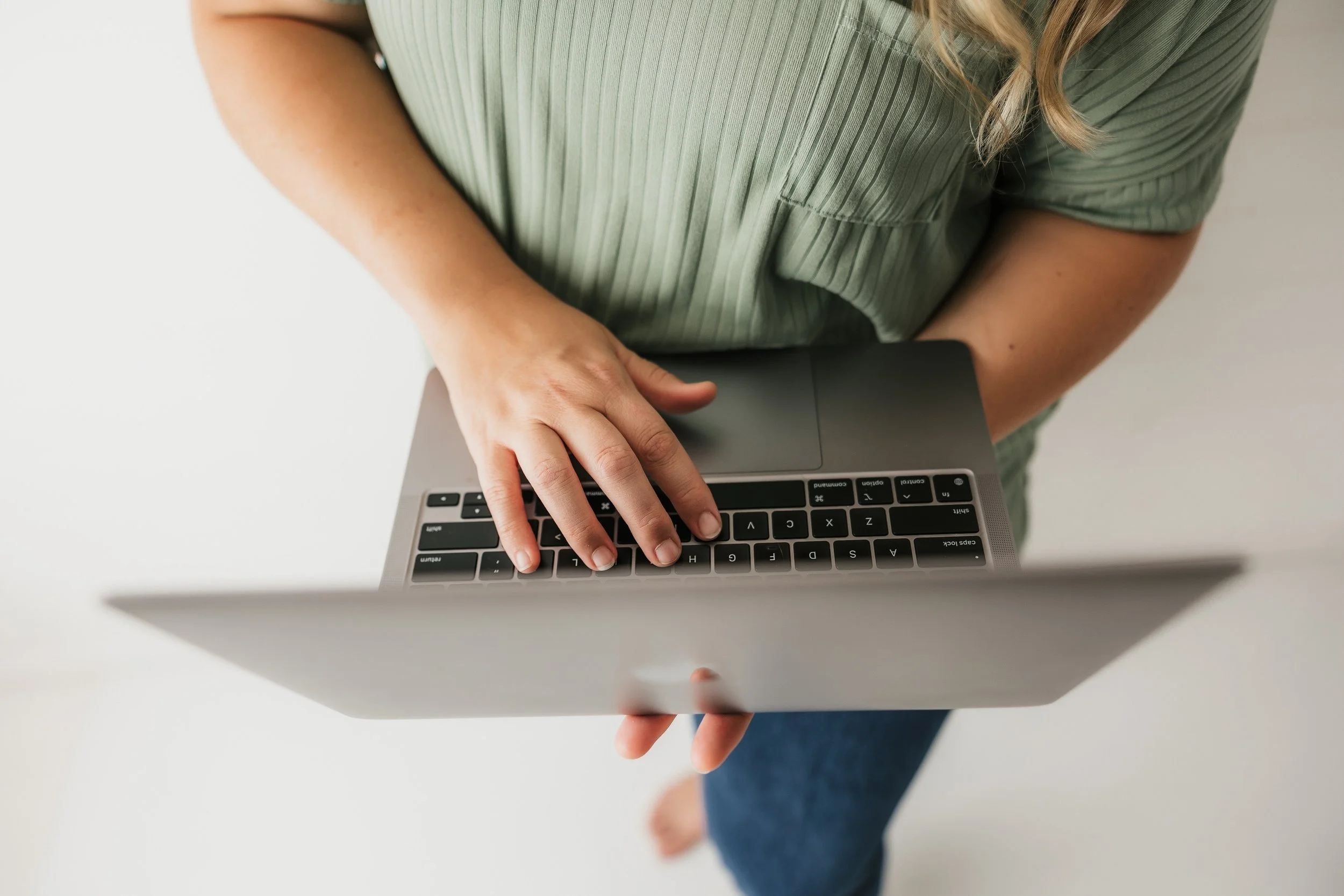 A person wearing a green, short-sleeved, ribbed shirt is using a laptop computer. The person's left hand is resting on the keyboard, and their right arm is bent with the hand near the laptop. The person is sitting on a blue chair or sofa with their legs crossed.