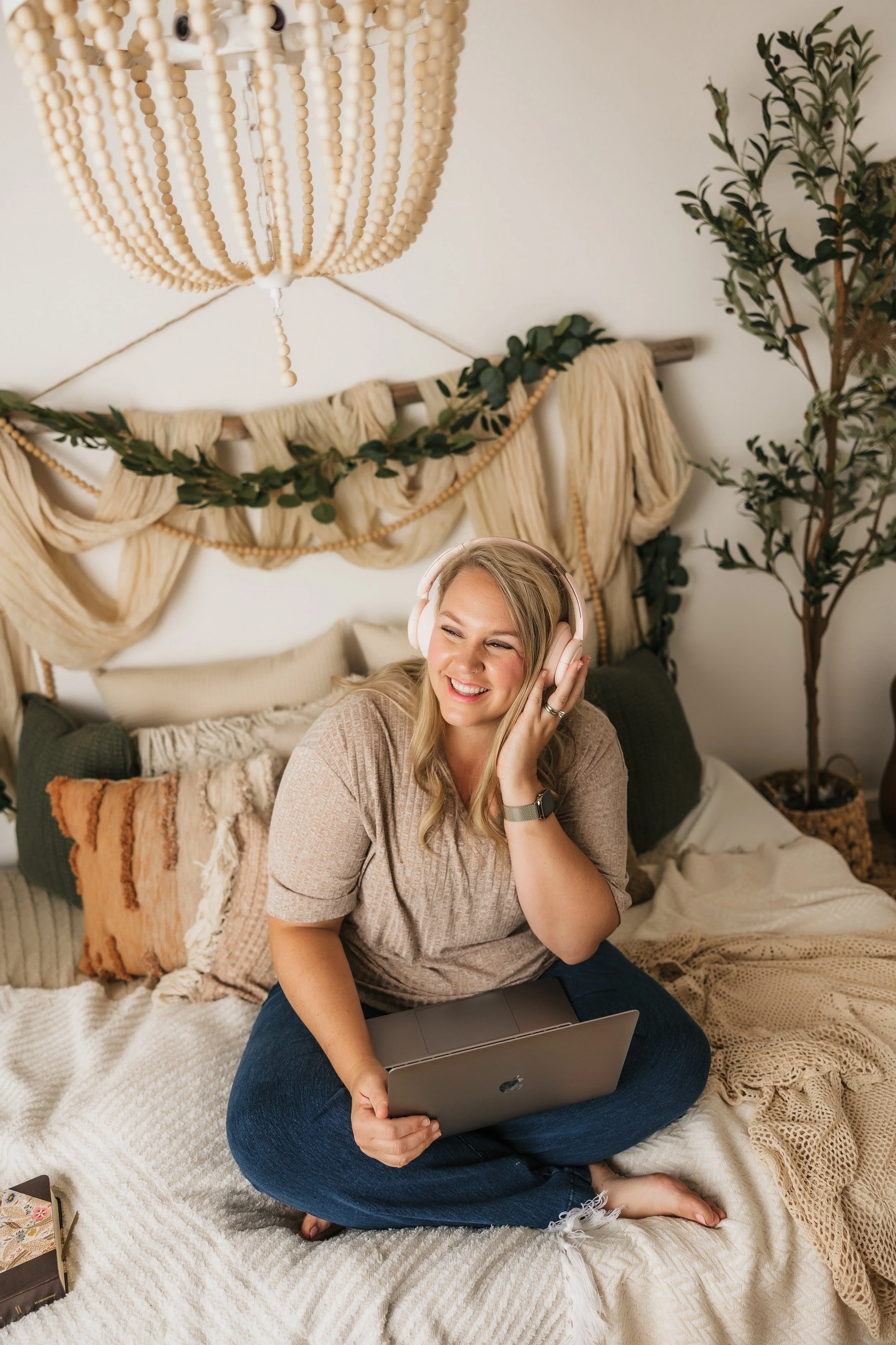 A smiling woman with blonde hair sitting cross-legged on a bed, wearing headphones, holding a silver laptop, with a cozy and bohemian bedroom with beige bedding, decorative pillows, a woven blanket, a macrame wall hanging, natural wood accents, and green plants in the background.