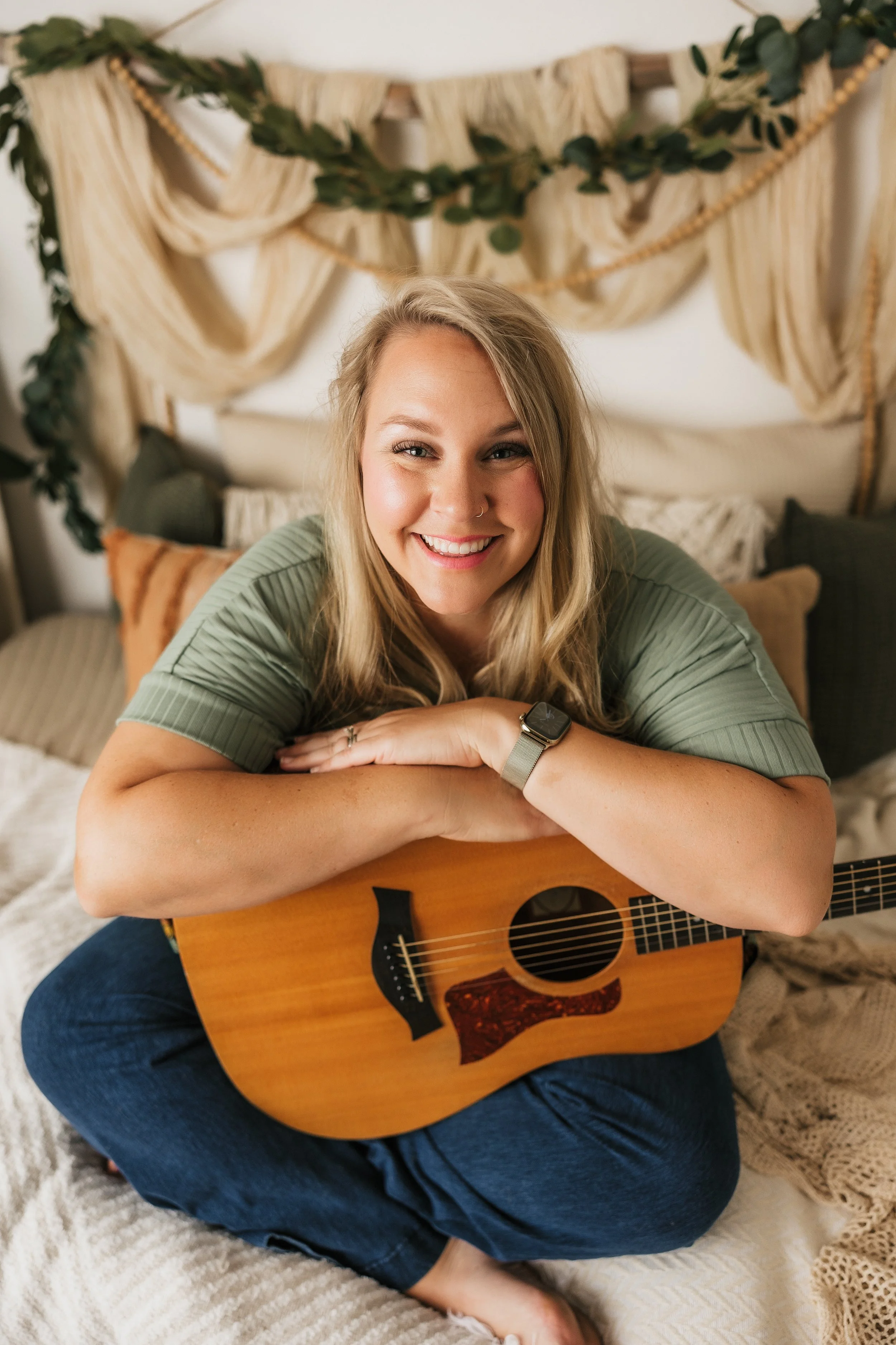 A young woman with blonde hair, smiling, sitting on a bed holding an acoustic guitar. Decor includes draped fabric, greenery, and cushions.