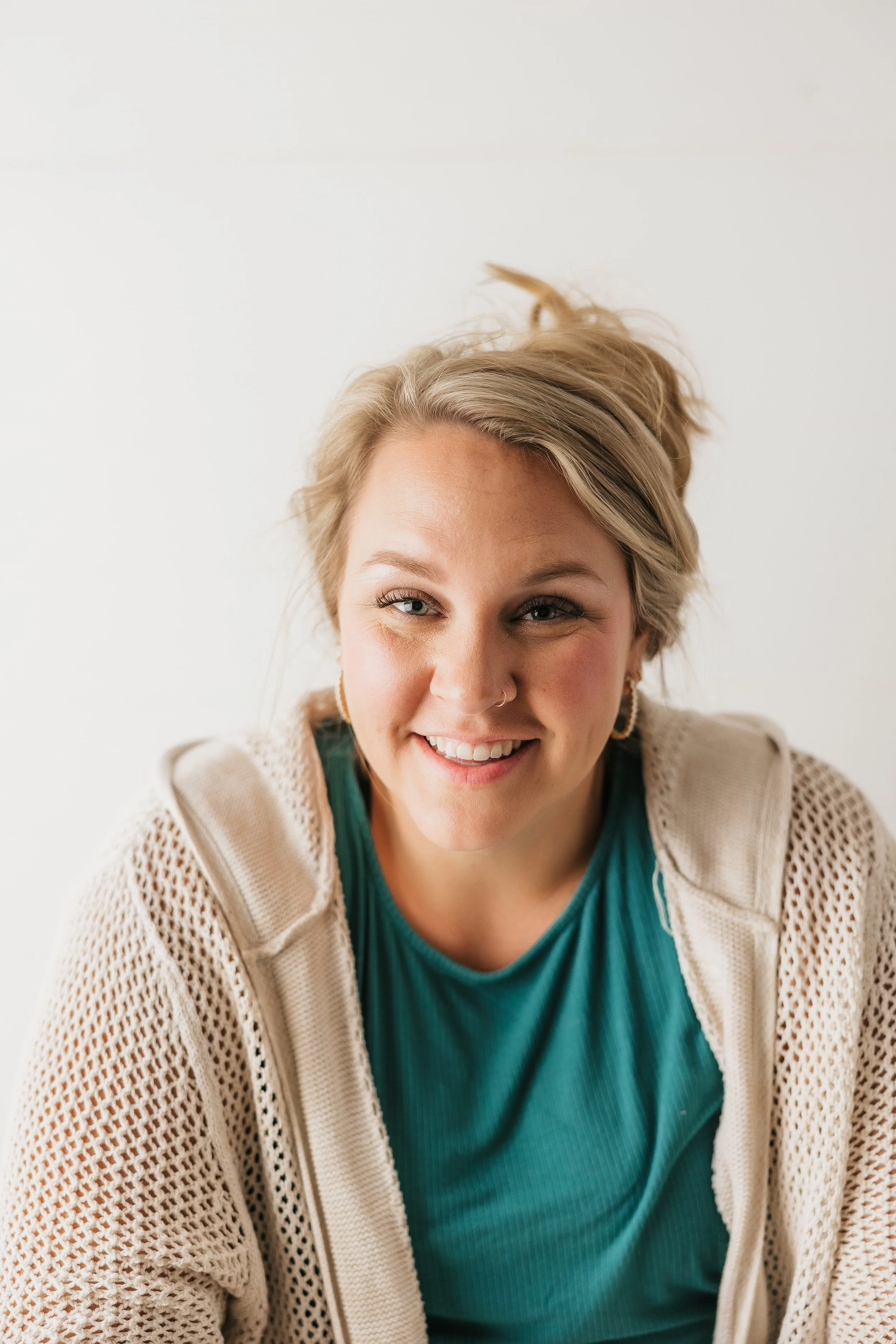 Close-up of a smiling woman with blonde hair, wearing a beige knit cardigan over a teal shirt, against a plain white background.