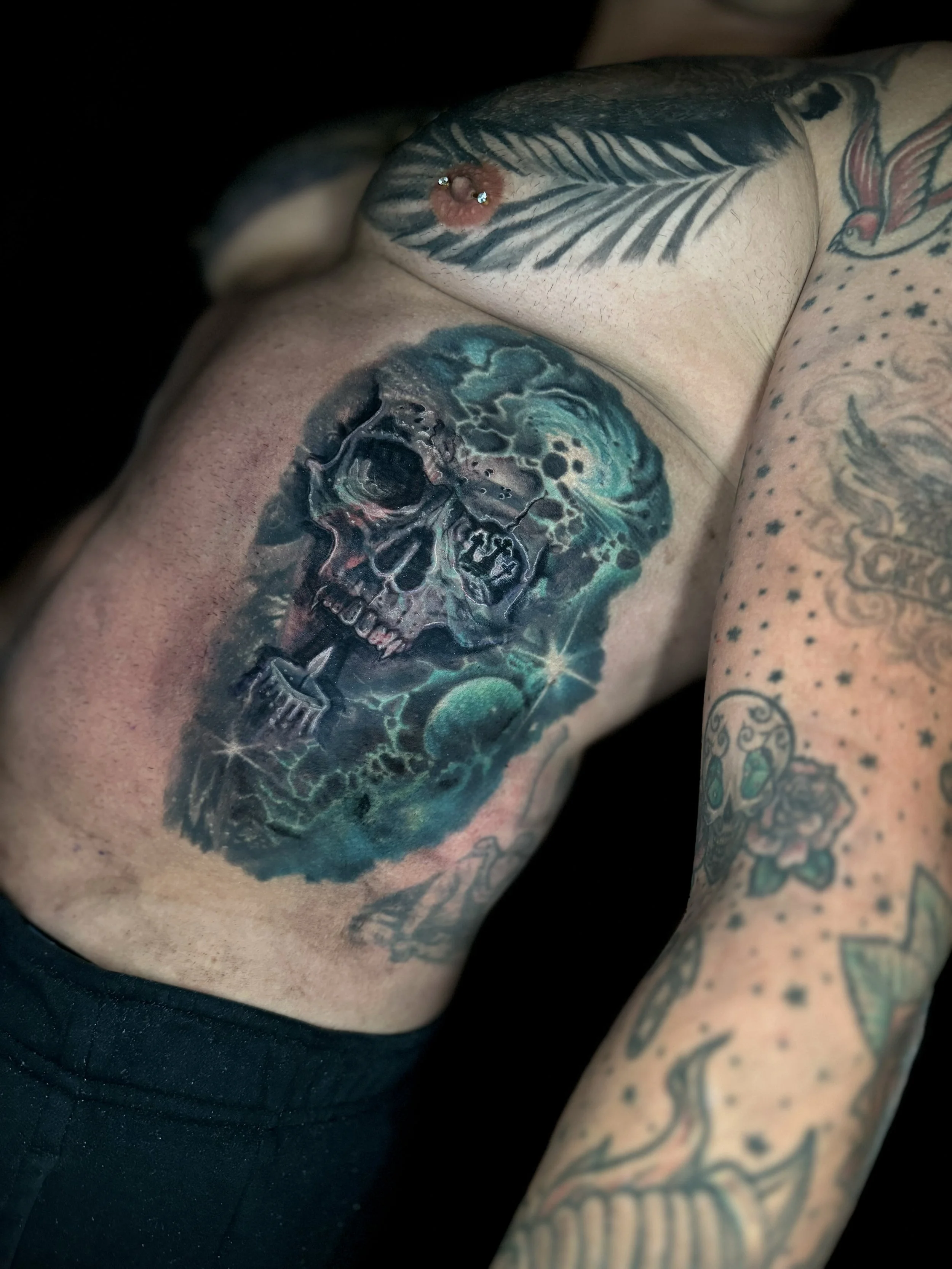 Close-up of a person's torso, showing a large colorful skull tattoo with vibrant shades of blue, green, black, and red, along with other smaller tattoos on the arm and chest.