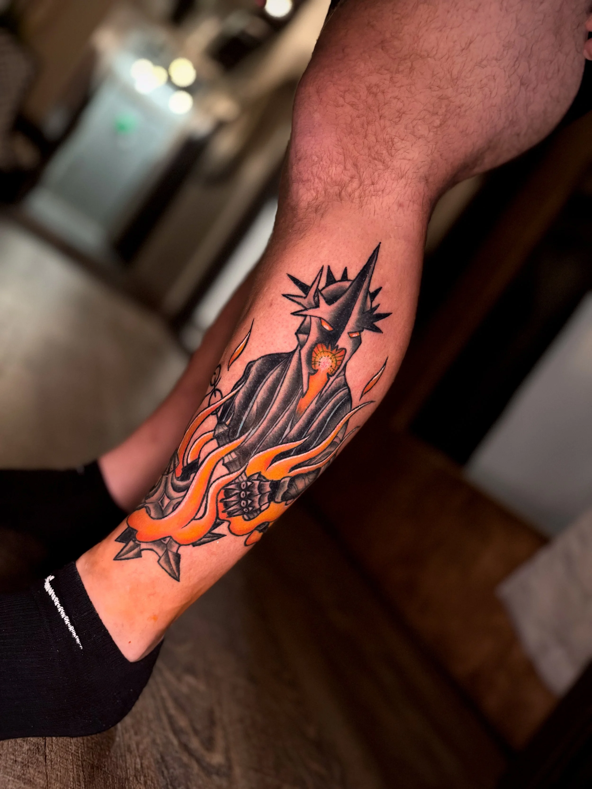 A person's arm with a colorful tattoo of a dark creature with orange eyes, sharp spikes, and flames.