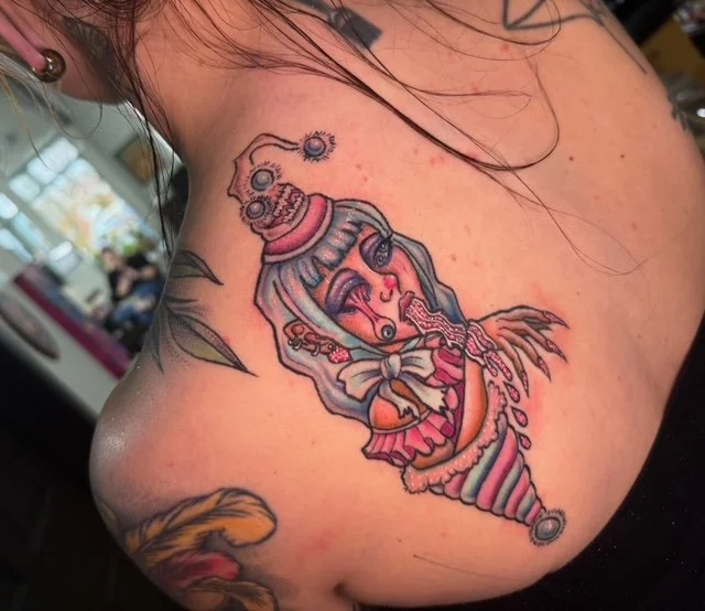 Tattoo of a surreal clown-like figure with colorful hair, large eyes, and a twisted expression, on a person's shoulder.
