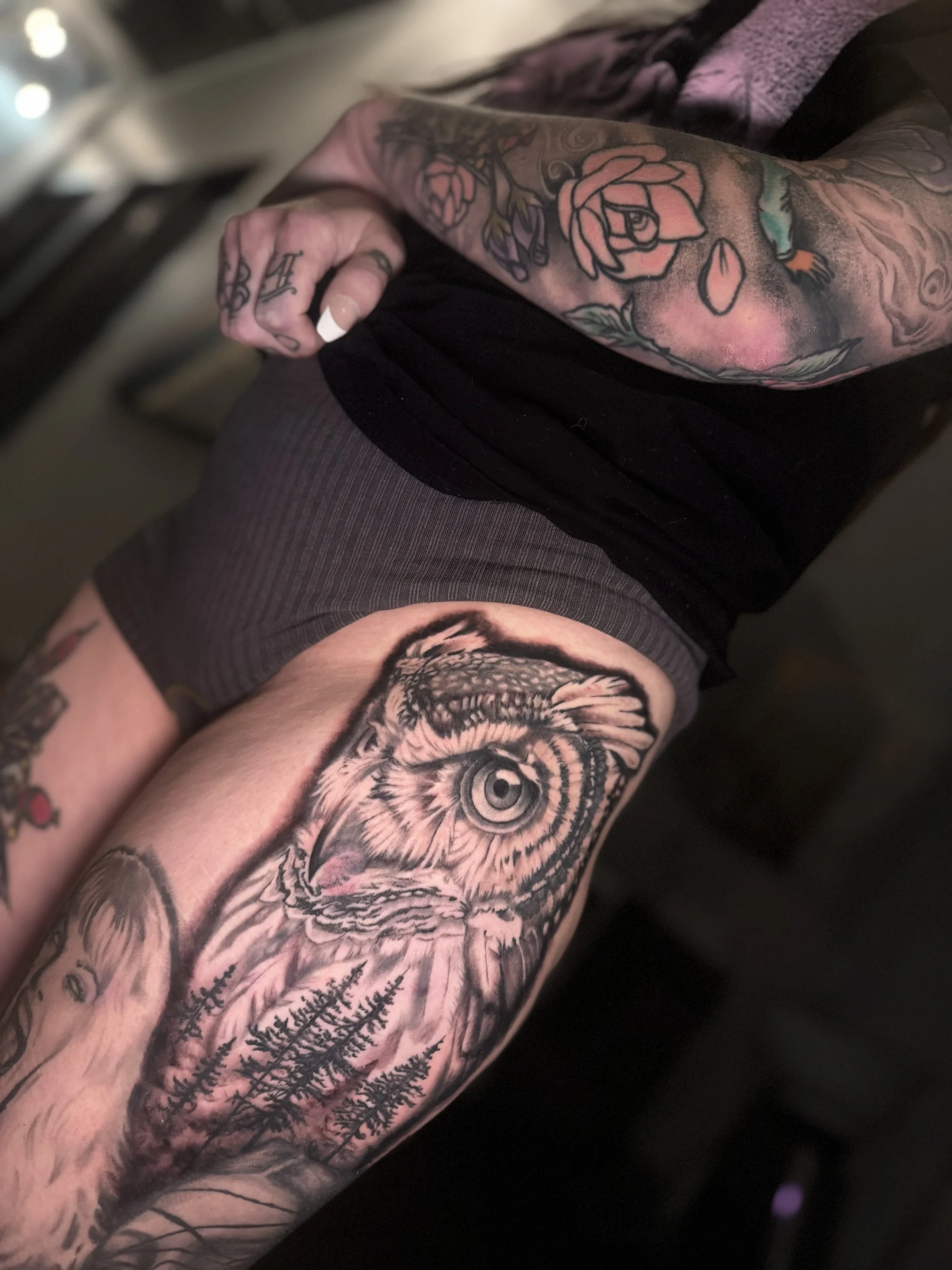 Close-up view of a tattooed arm with a large owl face on the lower arm and a rose tattoo above it, wearing a black shirt with a purple garment underneath.