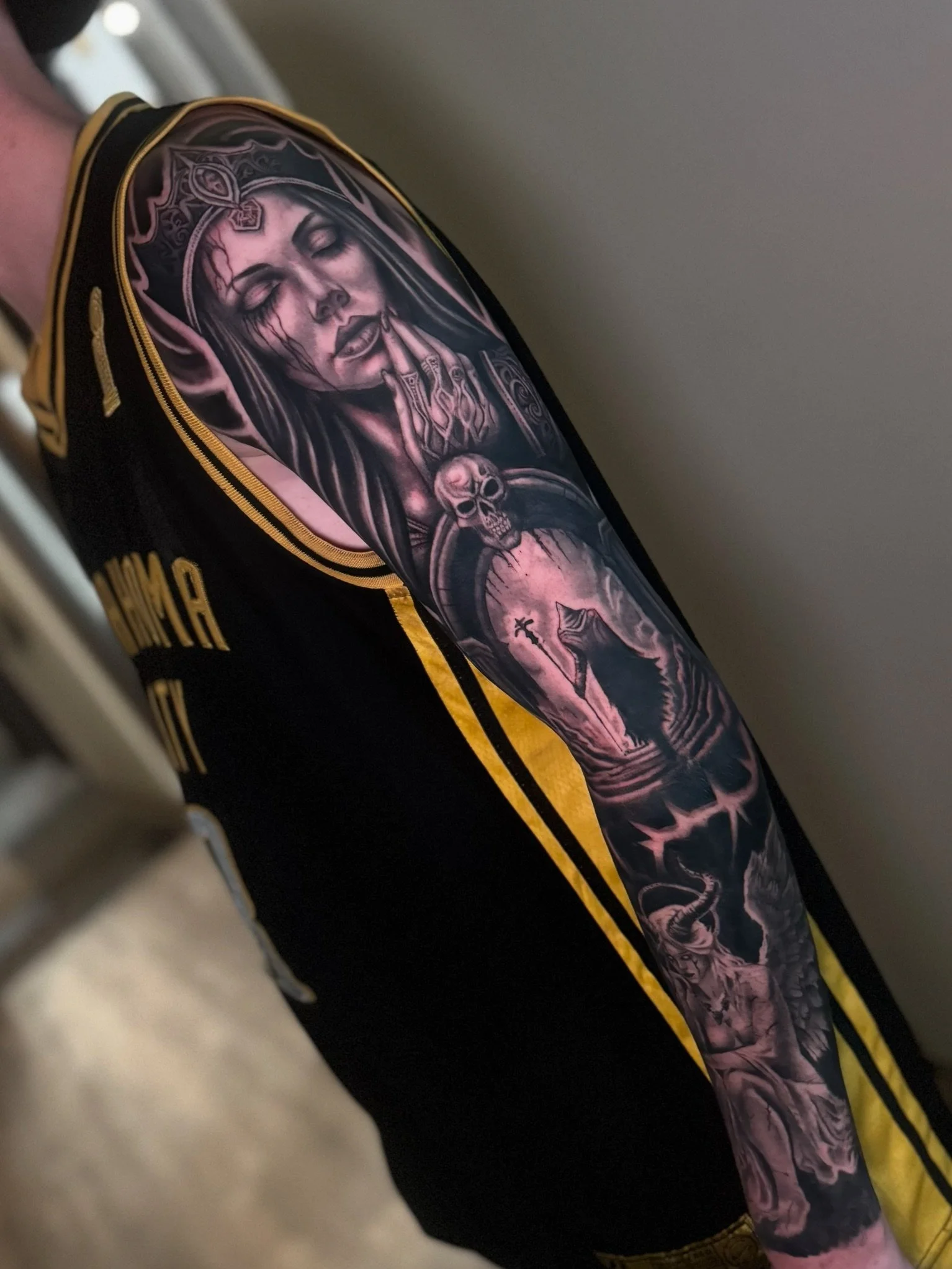 Close-up view of a tattoo on a person's arm, depicting a woman with closed eyes, a headdress, prayer hands, and skull elements, with black and gray ink details.