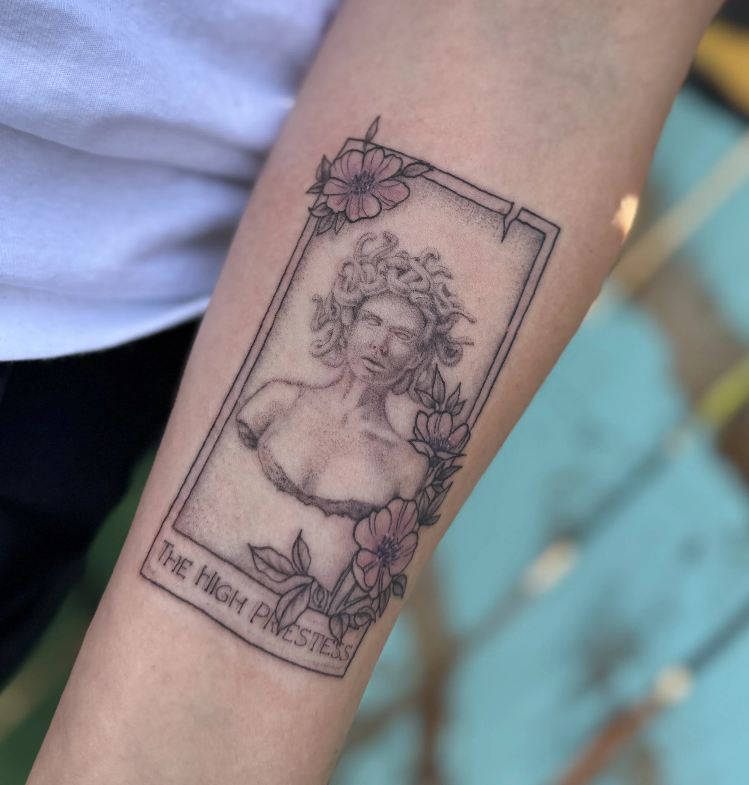 A tattoo of a Medusa head inside a rectangular frame on a person's forearm. The head is surrounded by pink flowers and greenery. There are words written below the frame, but they are partially obscured and not fully readable.