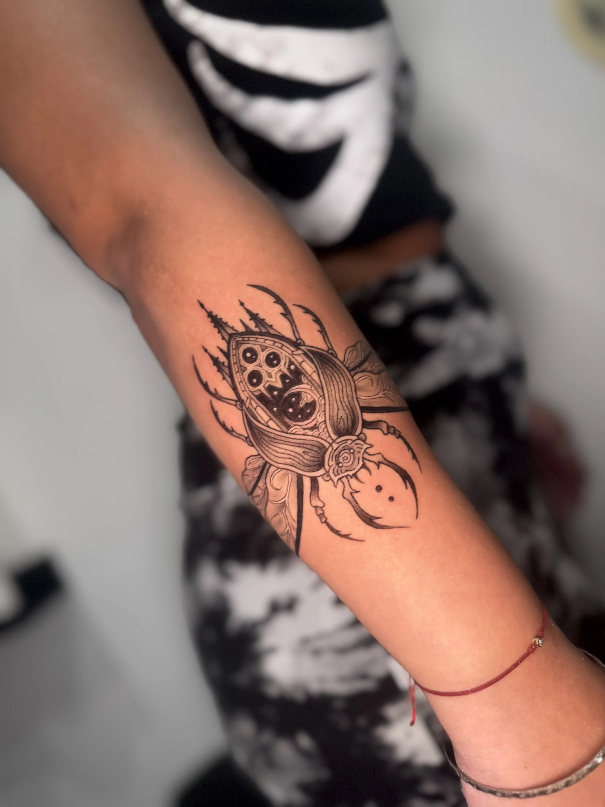 A person's arm with a tattoo of a beetle with detailed wings and antennae. The person is wearing a black and white striped shirt and has a red string bracelet on their wrist.