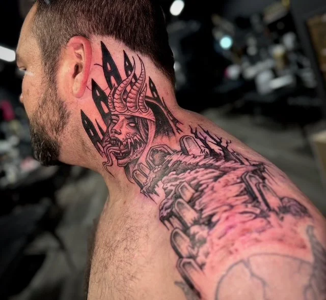 Tattoo of a demonic face with horns and flames on a person's neck and shoulder