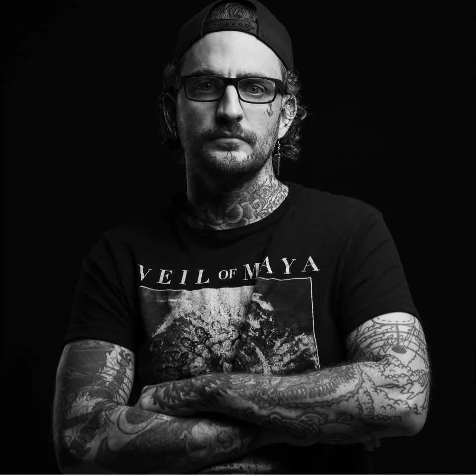 A man with glasses, tattoos, and a backward cap, wearing a Veil of Maya T-shirt, poses against a black background.