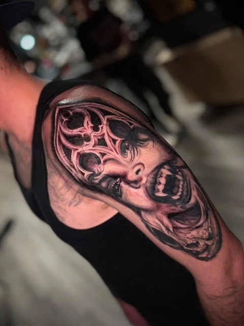 Tattoo of a screaming skull with a decorative circular pattern above it on person's upper arm.