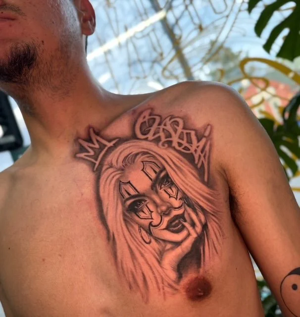 Tattoo of a woman with long hair, facial markings, and a crown on her chest, with graffiti-style text above her.