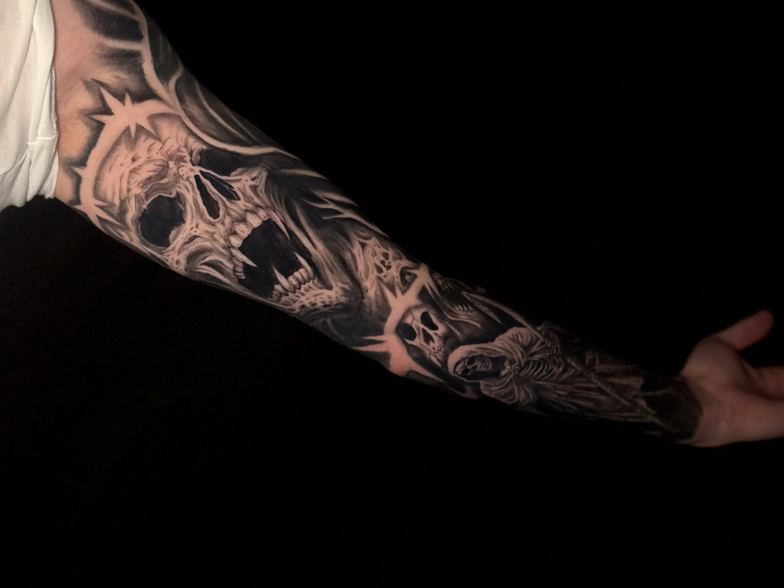 A person's arm covered in black and grey tattoos, including a skull and various designs, set against a dark background.