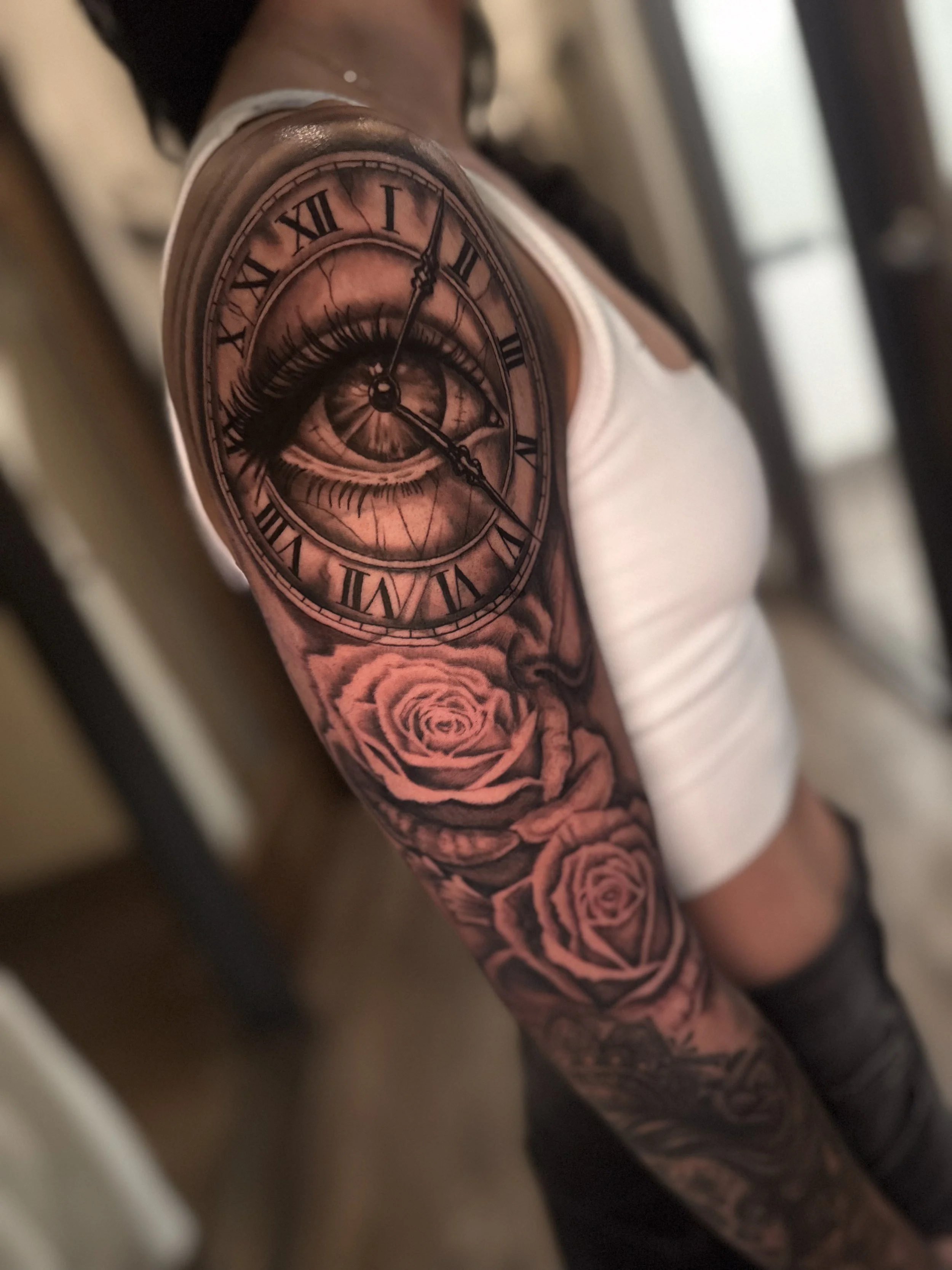 Tattoo of a clock with Roman numerals, an eye, and roses on a person's arm.