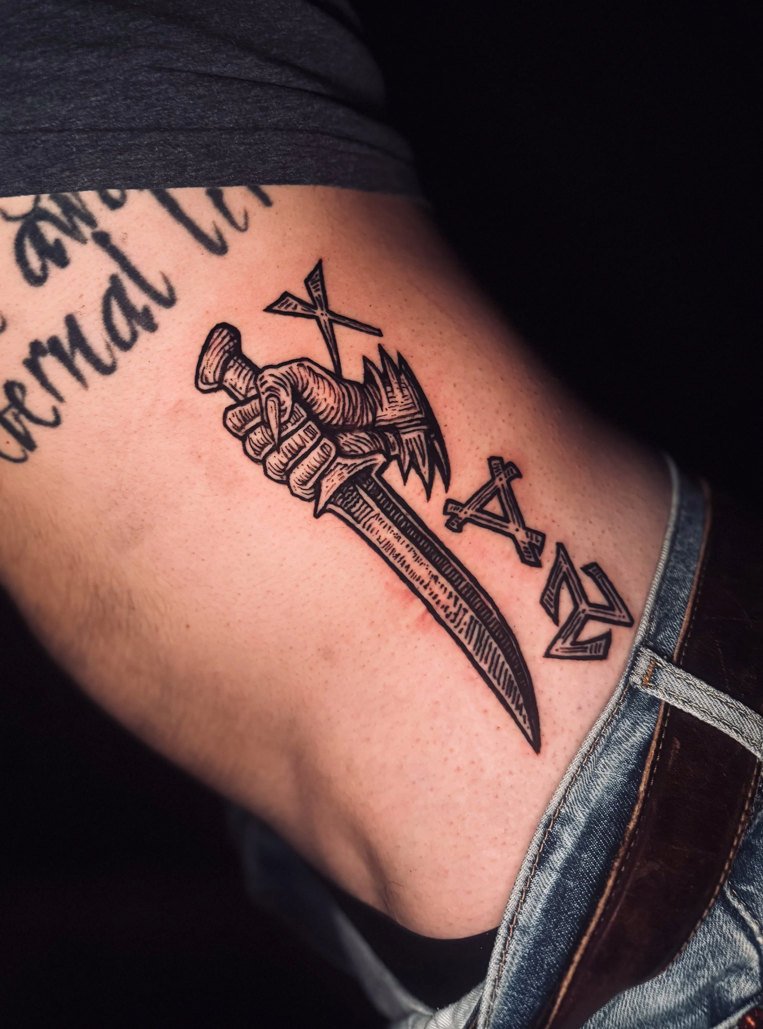 Close-up of a tattoo on a person's upper arm, depicting a hand gripping a sword with an arrow through it; part of text is visible.