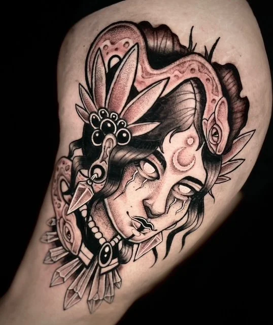 Tattoo of a woman's face with a snake wrapped around her head, surrounded by flowers and jewelry, in black and gray ink.