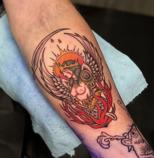 Tattoo of a woman with angel wings, wearing a crown, surrounded by flames on a forearm.