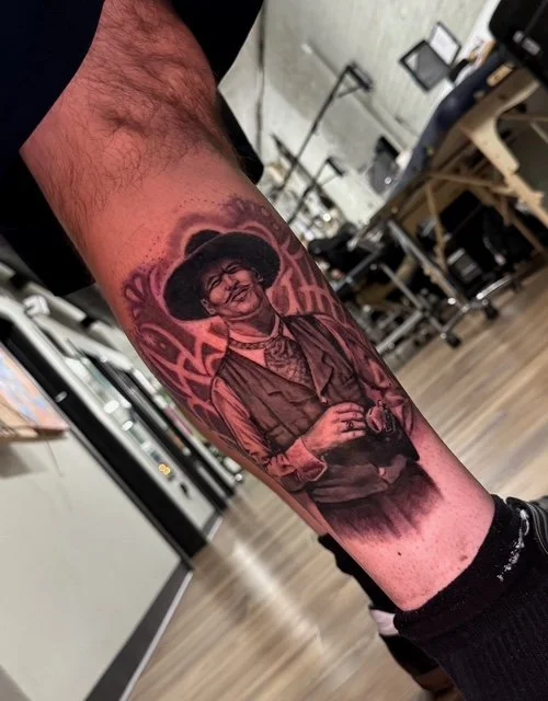 A tattoo on a person's forearm depicting a woman with curly hair, dressed in vintage clothing, smiling and holding a camera. The background includes decorative elements and text.