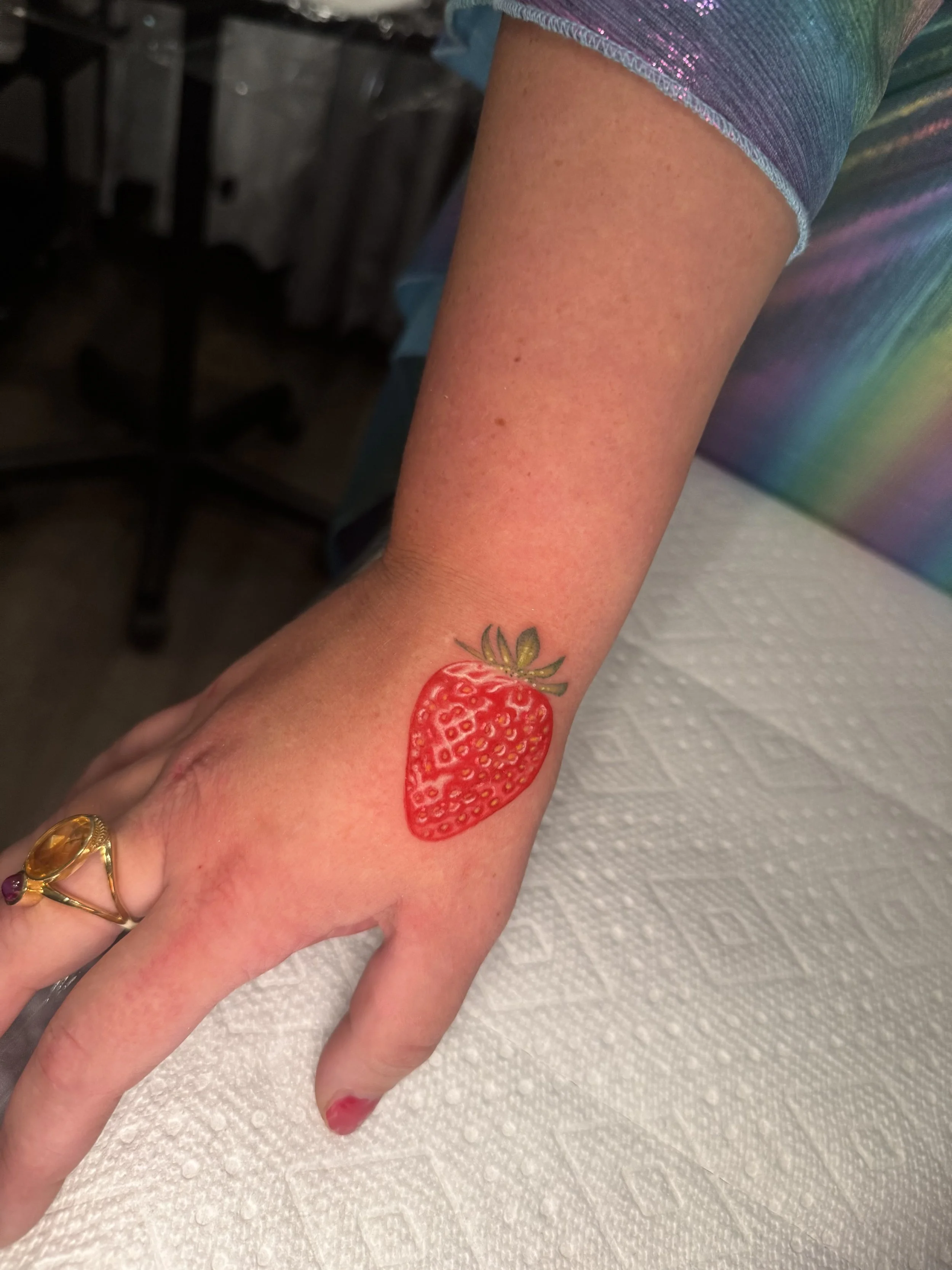 A person's forearm with a strawberry tattoo on the wrist, resting on a white textured surface with colorful clothing in the background.