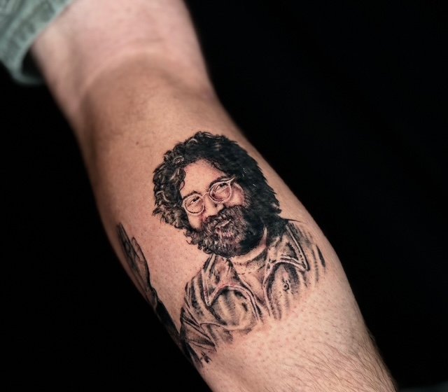 Tattoo of a man with curly hair, glasses, and a beard, wearing a jacket on a person's arm.
