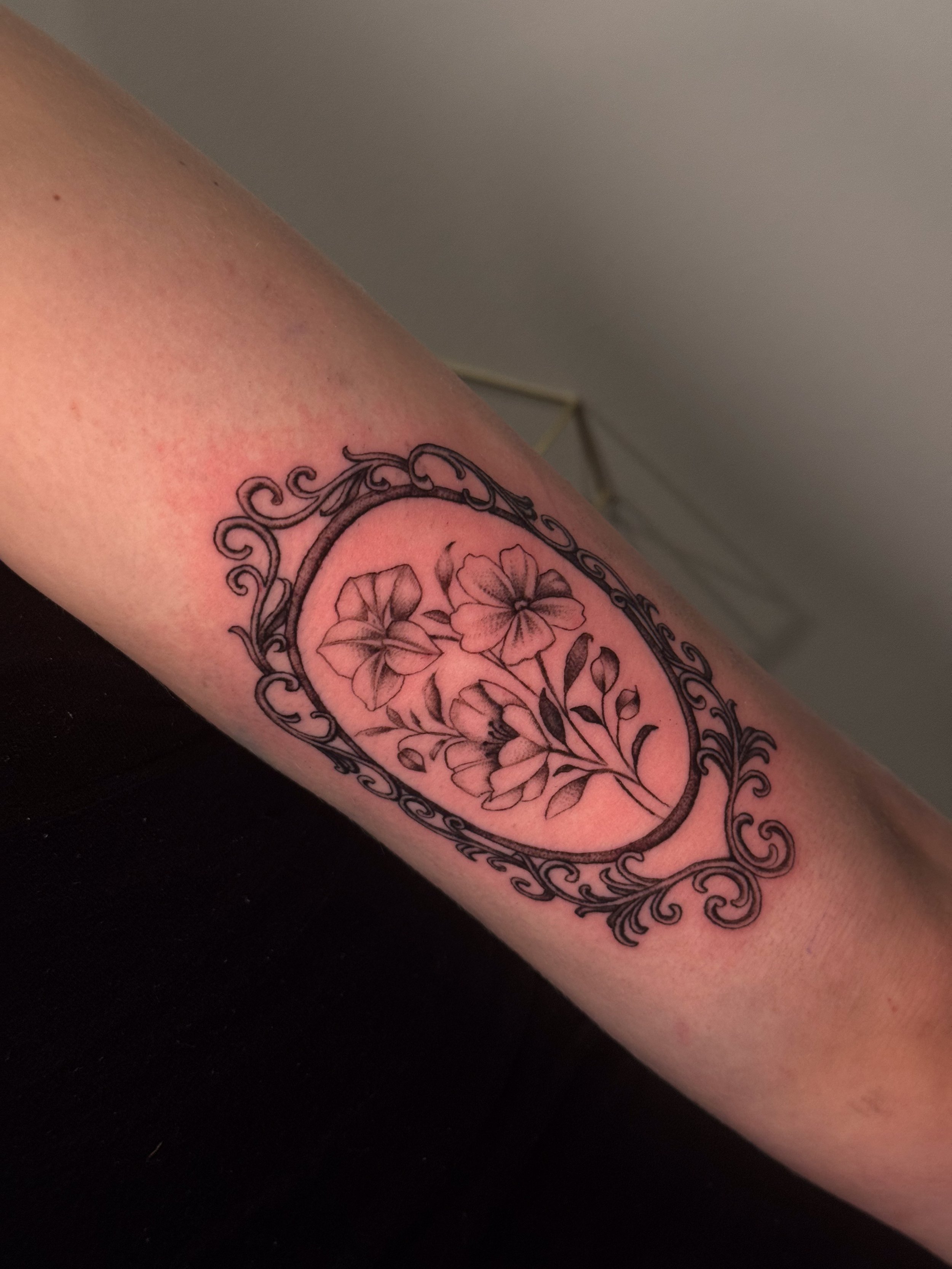 A tattoo of flowers in a decorative oval frame on a person's arm.