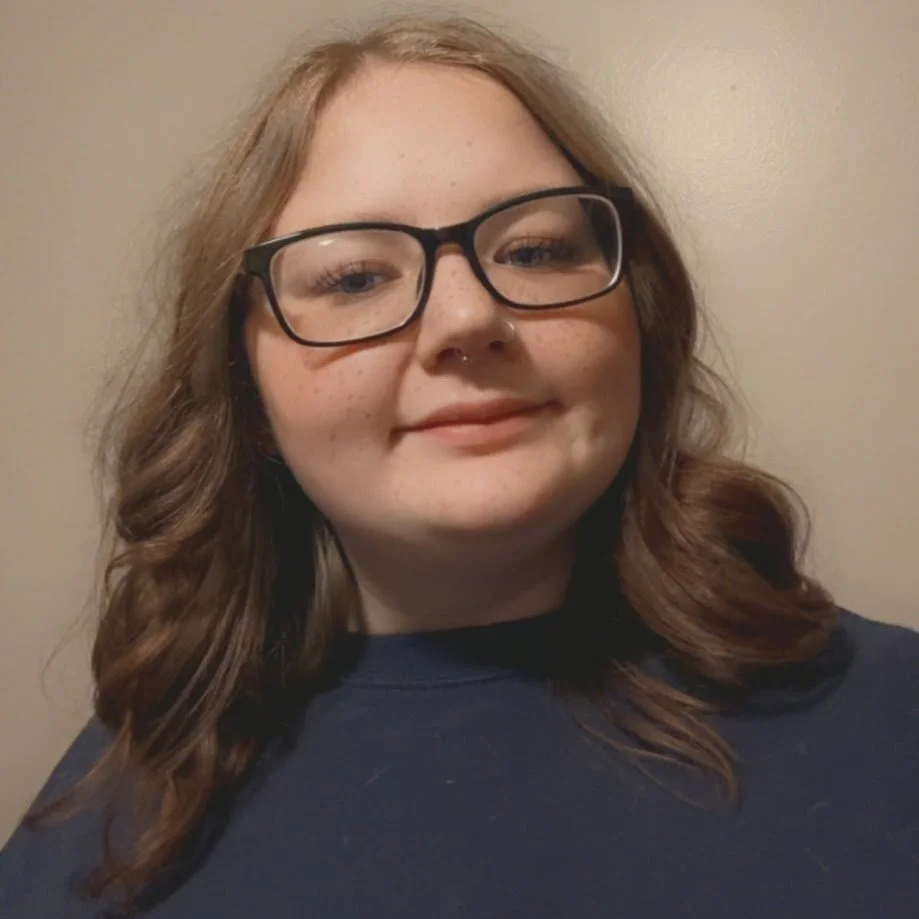 A young woman with wavy brown hair, glasses, and a septum piercing, smiling against a plain beige wall.