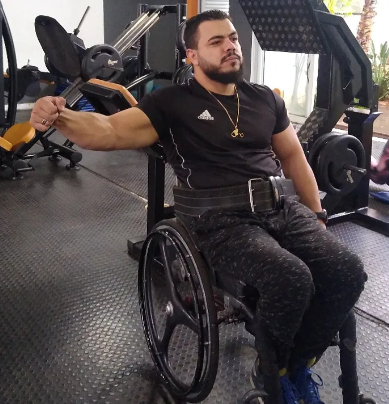 A man in a wheelchair at a gym, flexing his right arm and looking to the side, dressed in black workout clothes, with gym equipment in the background.