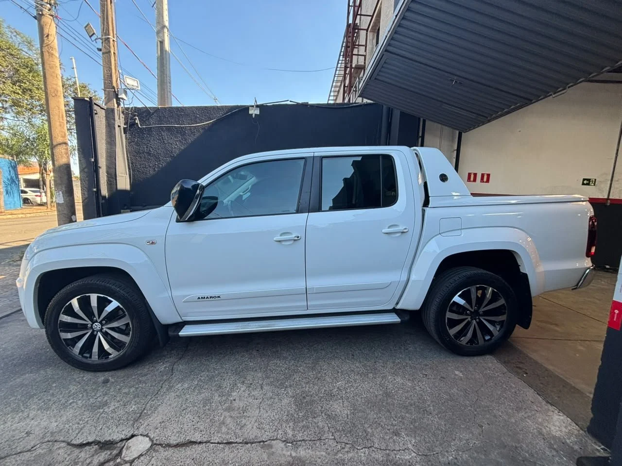 A white Volkswagen Amarok pickup truck parked outdoors next to a black wall and under a metal awning. The truck has black and silver alloy wheels, tinted windows, and a canopy over the truck bed.