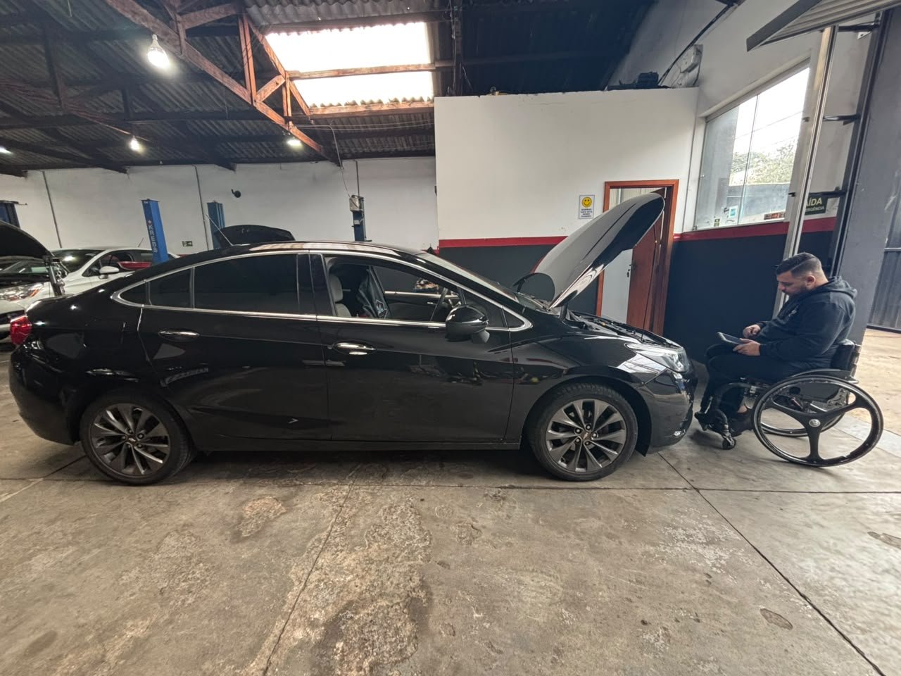 A black sedan with its hood open parked inside a workshop. A man in a wheelchair is sitting next to the car, looking at a tablet or device.