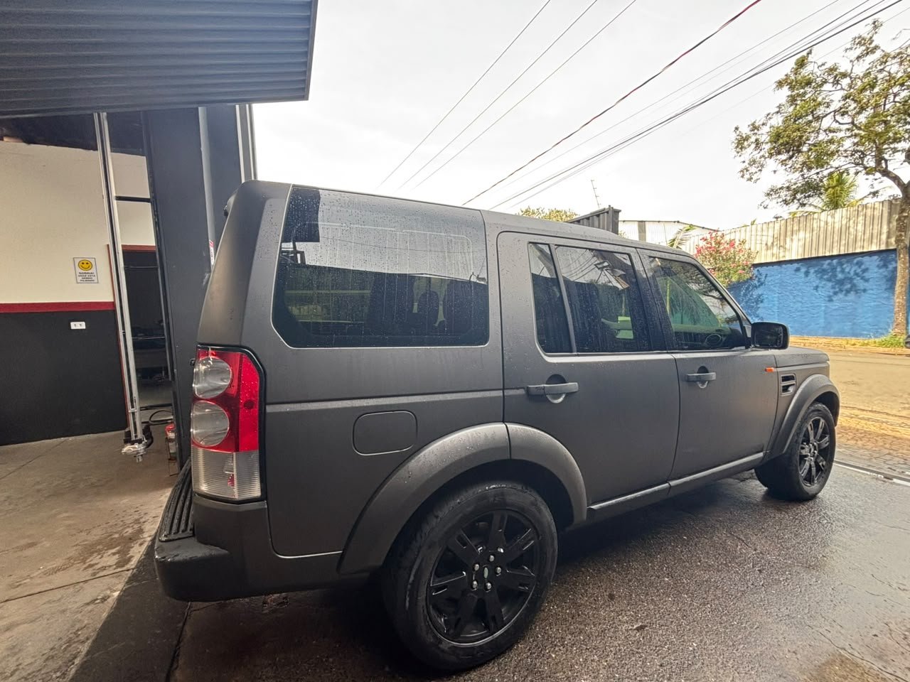 Gray SUV parked outside a building with black tires and tinted windows, rain droplets on the vehicle, and a colorful background with trees and a blue wall.