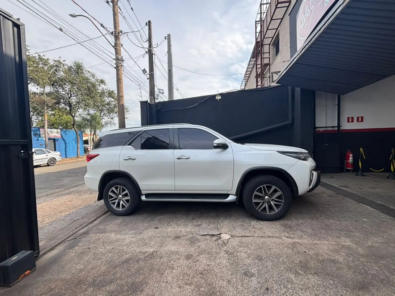 White SUV parked outside a building with a dark blue wall, utility poles, and trees in the background.